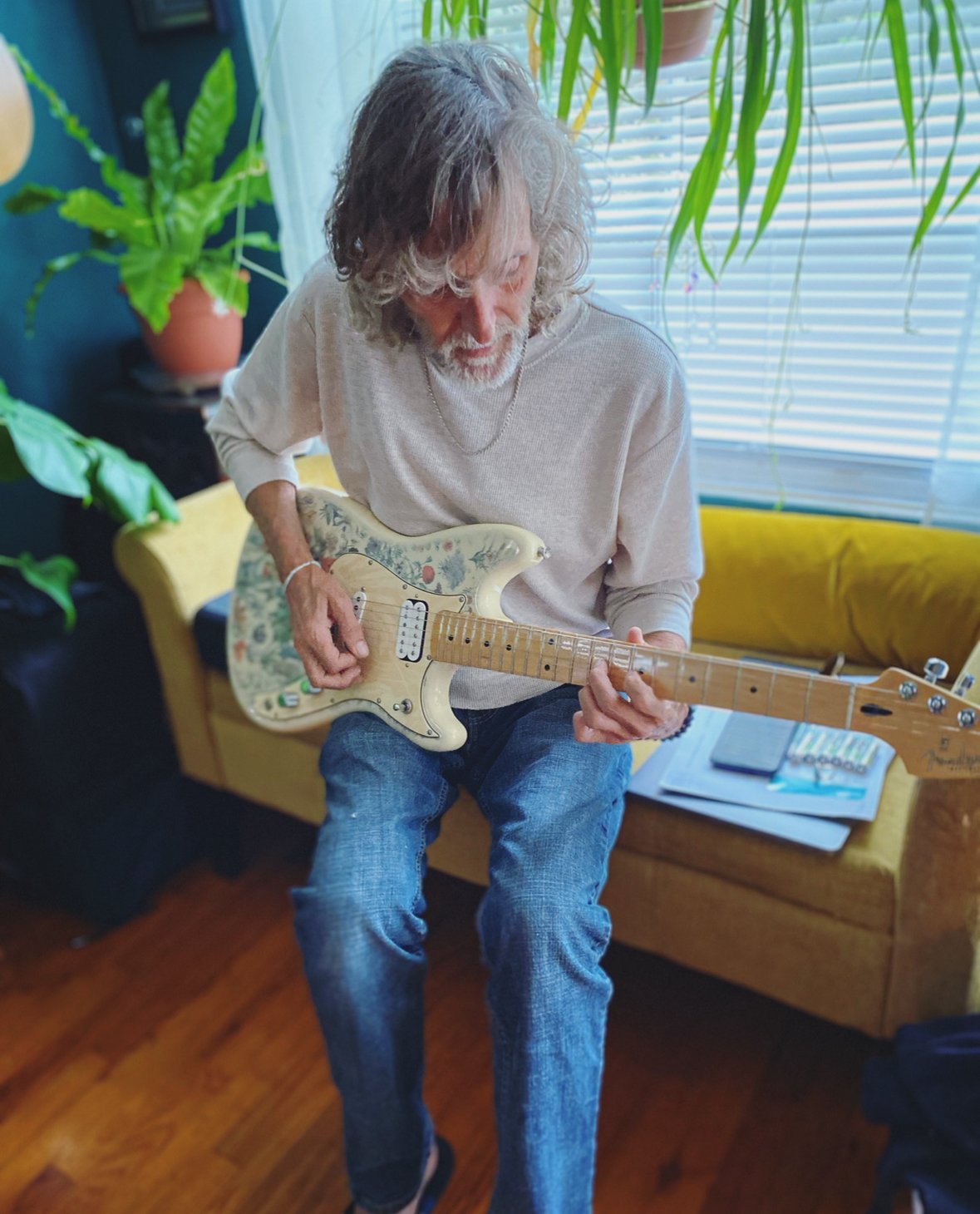 A man with gray hair and a beard playing a vintage Fender Stratocaster electric guitar in a cozy room with plants and a yellow sofa.