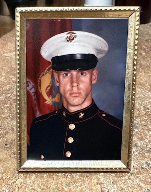 A framed portrait of a young Marine in dress uniform with a white cover and black brim, against a backdrop featuring the U.S. Marine Corps emblem.