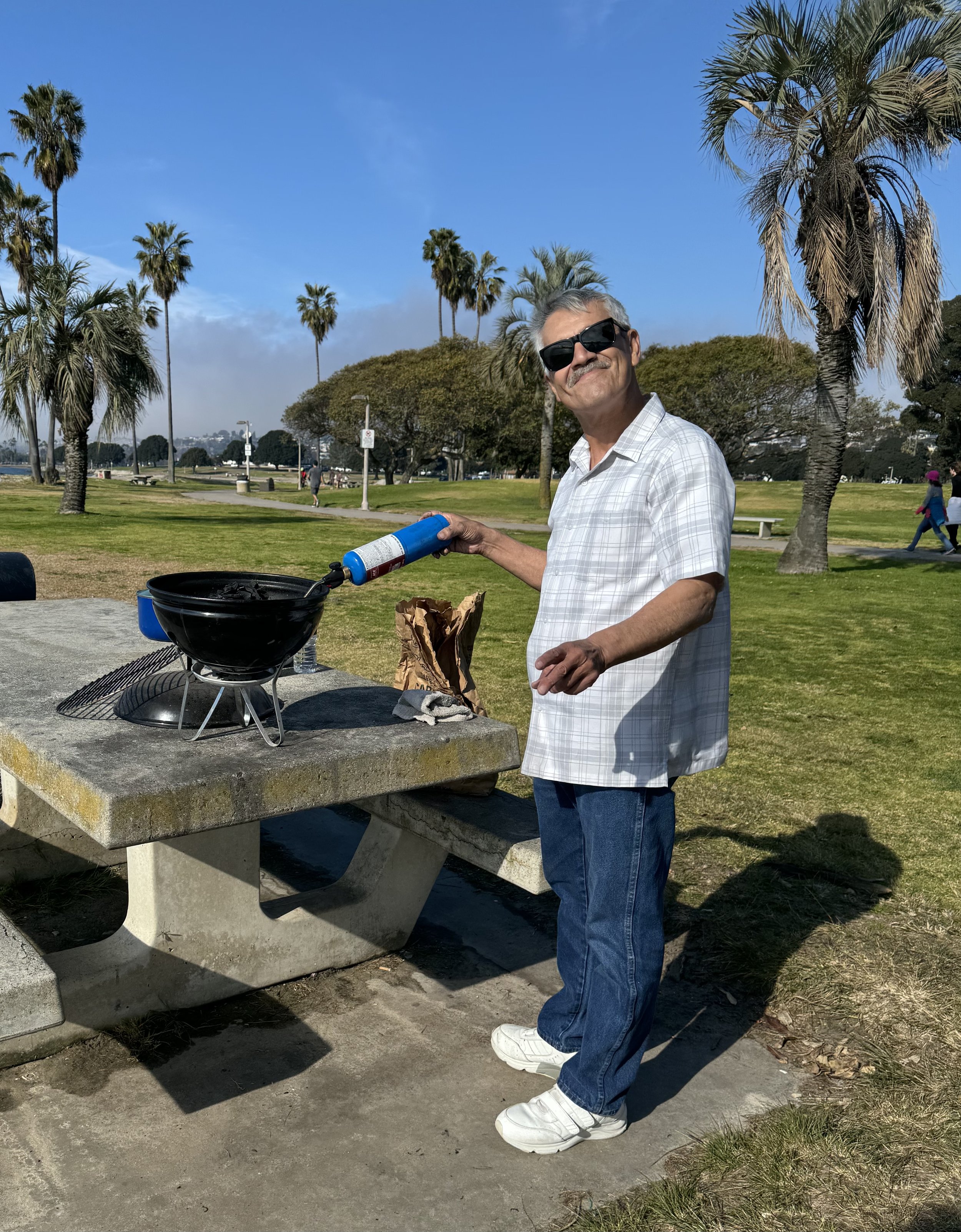 A man wearing sunglasses and a white plaid shirt standing at an outdoor picnic table, pouring lighter fluid into a black tabletop grill in a park with palm trees and a clear blue sky.