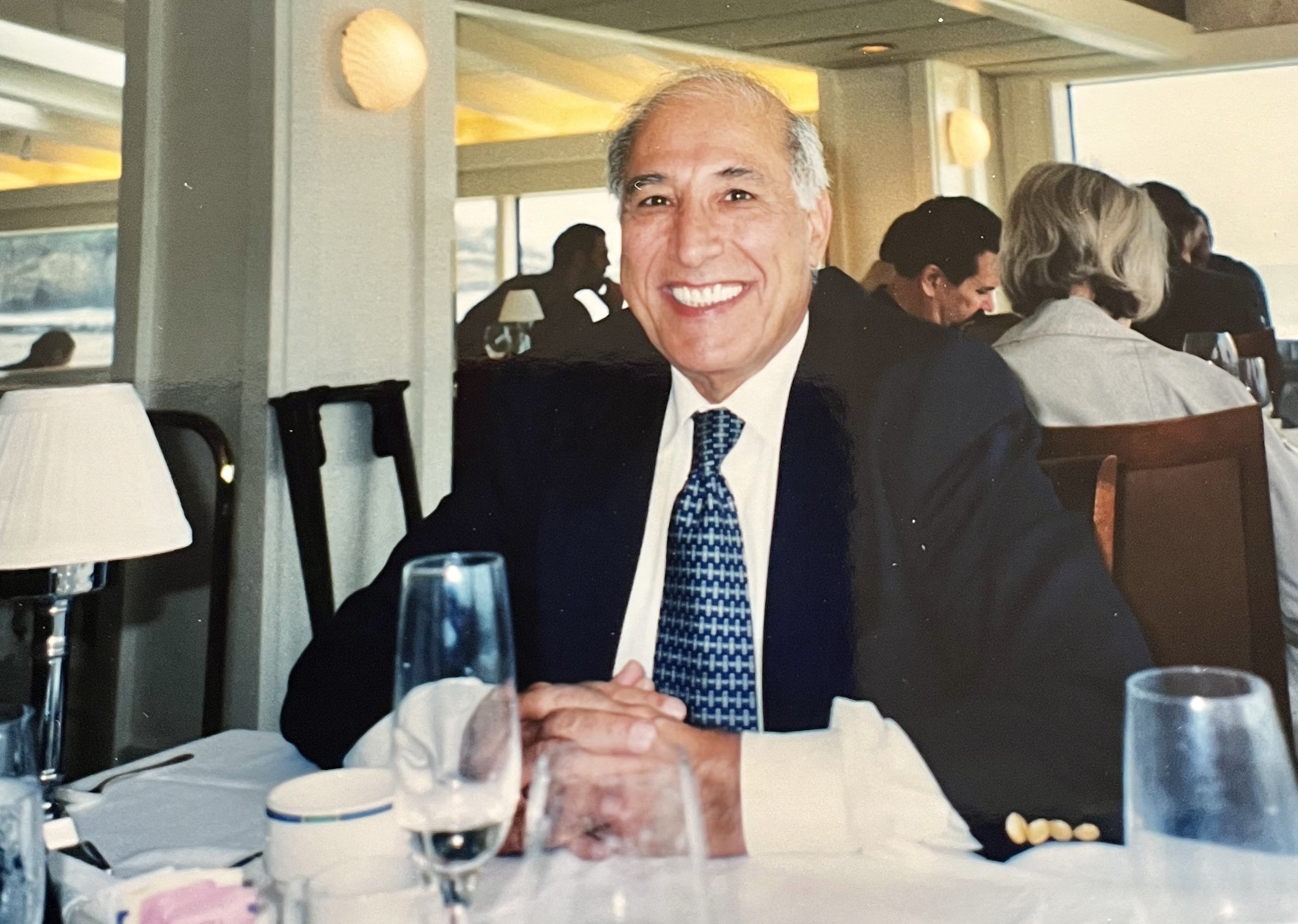 A smiling middle-aged man in a black suit, white shirt, and patterned blue tie sitting at a restaurant table, with glasses, a lamp, and some tableware, during daytime at a waterfront restaurant.
