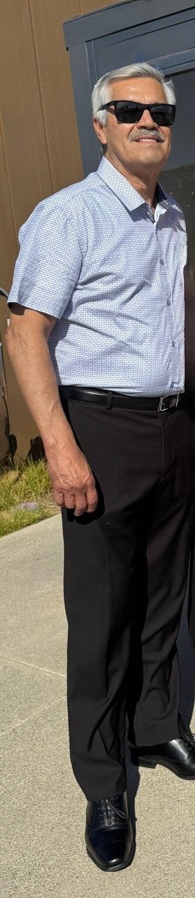 Man with gray hair, wearing sunglasses, a light blue short-sleeve button-up shirt, black pants, and black shoes, standing outdoors near a building.