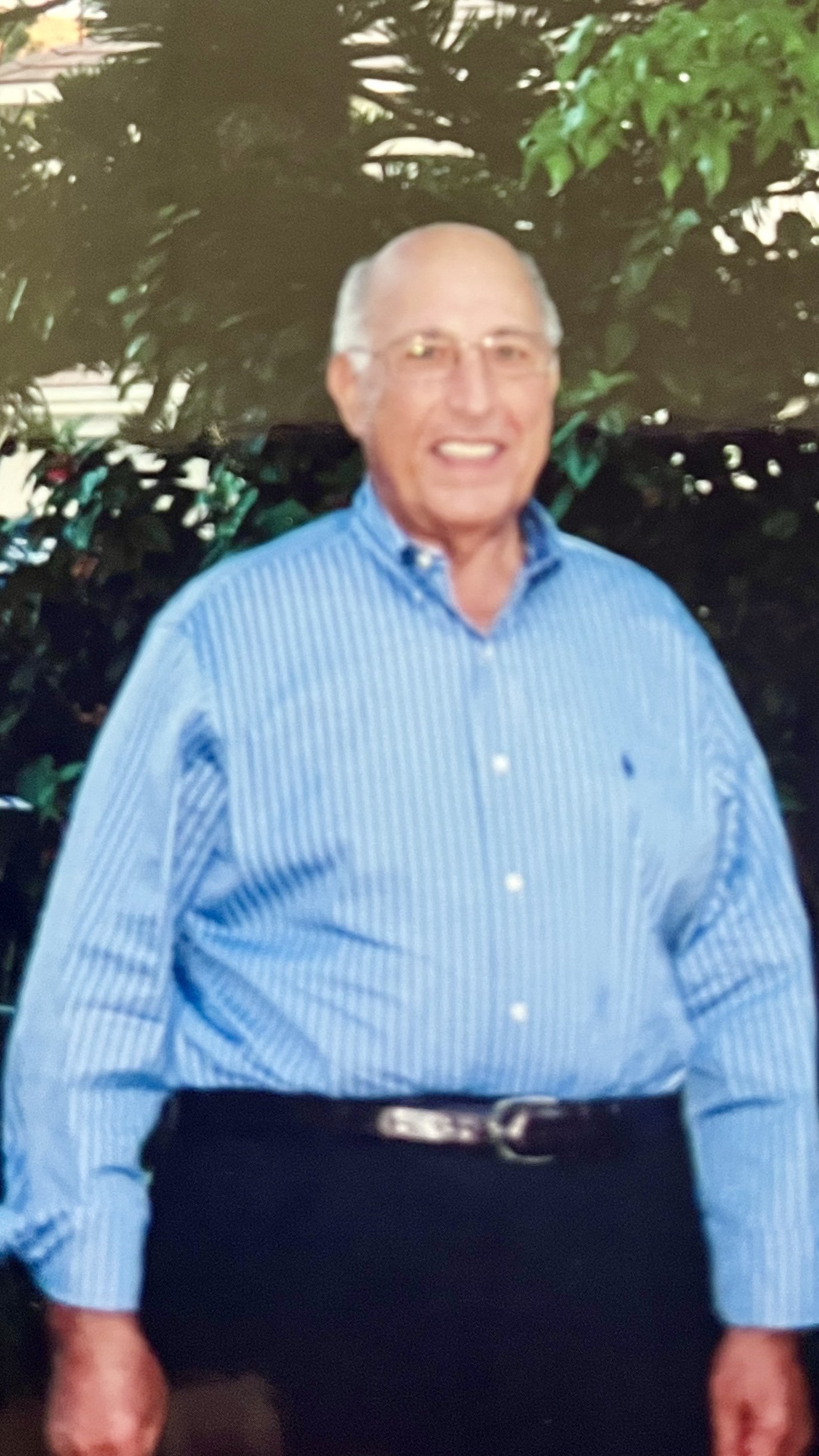 Smiling elderly man in a blue striped dress shirt with glasses, standing outdoors with green foliage in the background.