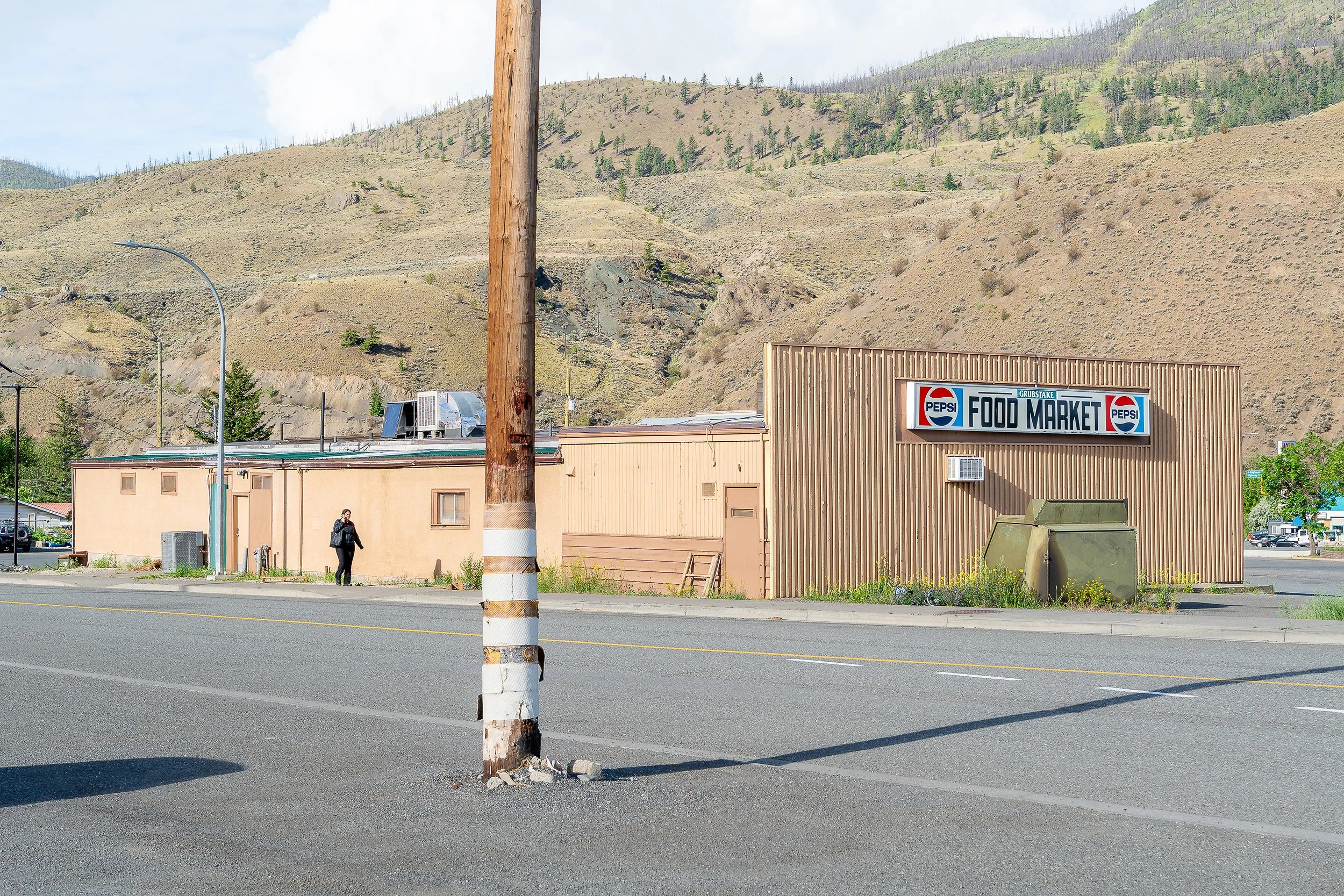 A street scene with a pedestrian walking near a beige food market building, a utility pole, and a mountain with sparse trees in the background.