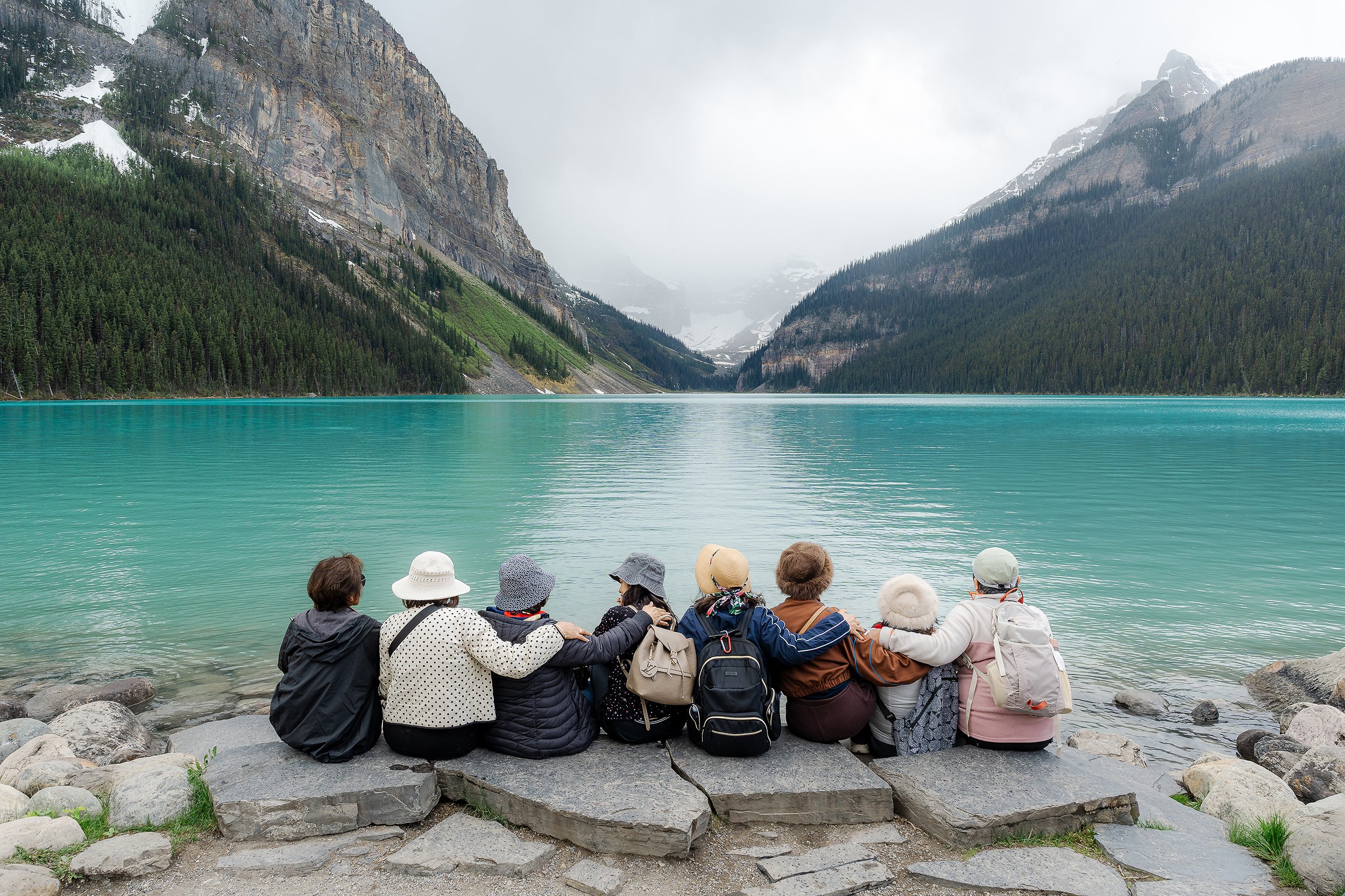 Group of people sitting on rocks by a turquoise lake surrounded by mountains and dense pine forests, on a cloudy day.