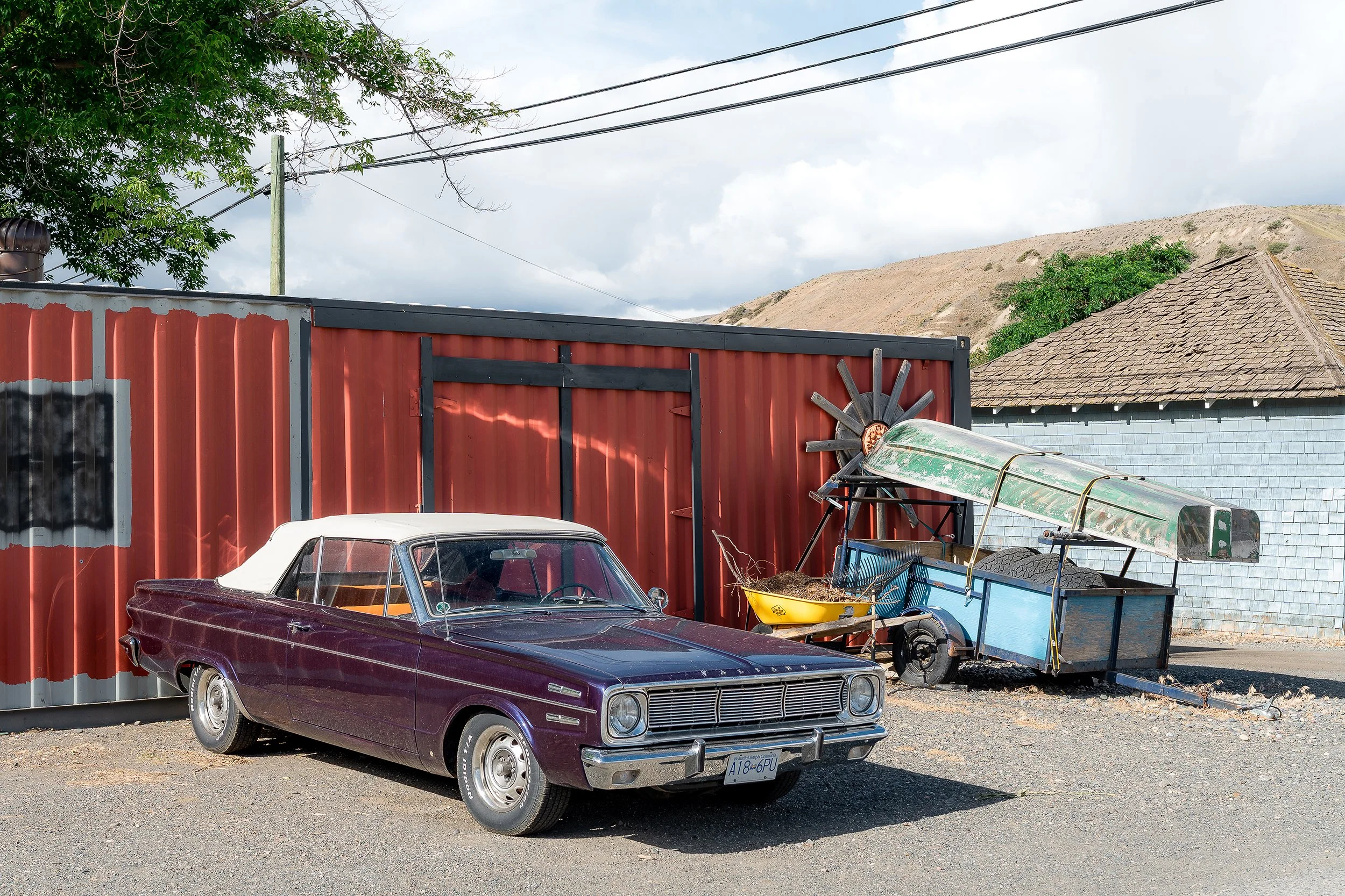 A vintage purple convertible car parked in front of a red corrugated metal fence. Behind the fence, there are other objects including a wheelbarrow, some outdoor equipment, and part of a building with gray brick siding and a sloped shingle roof. In t