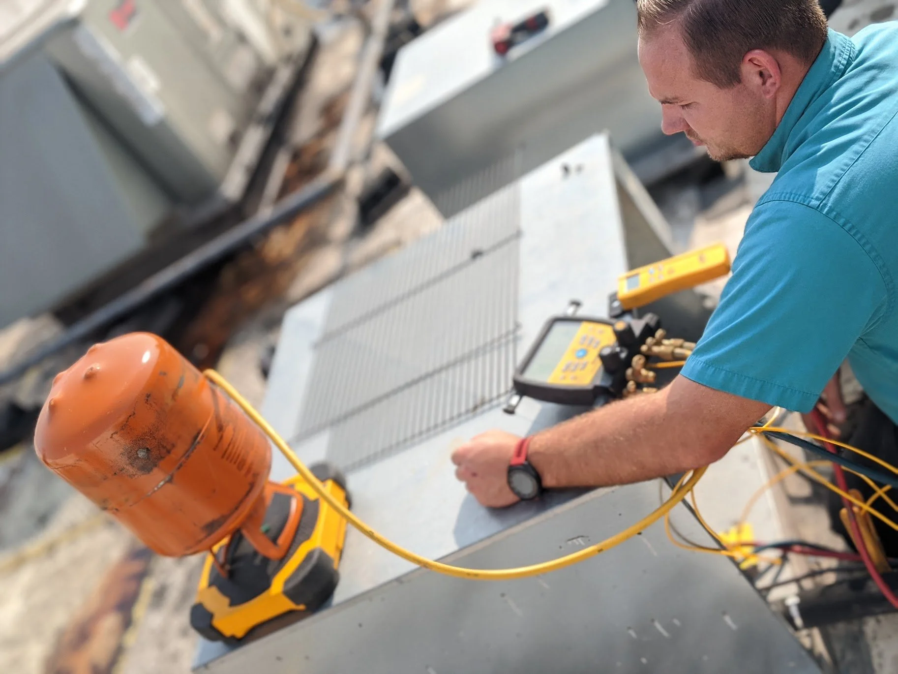 A man in a blue shirt working on an HVAC or refrigeration unit using a digital multimeter and tools on a rooftop or industrial setting.