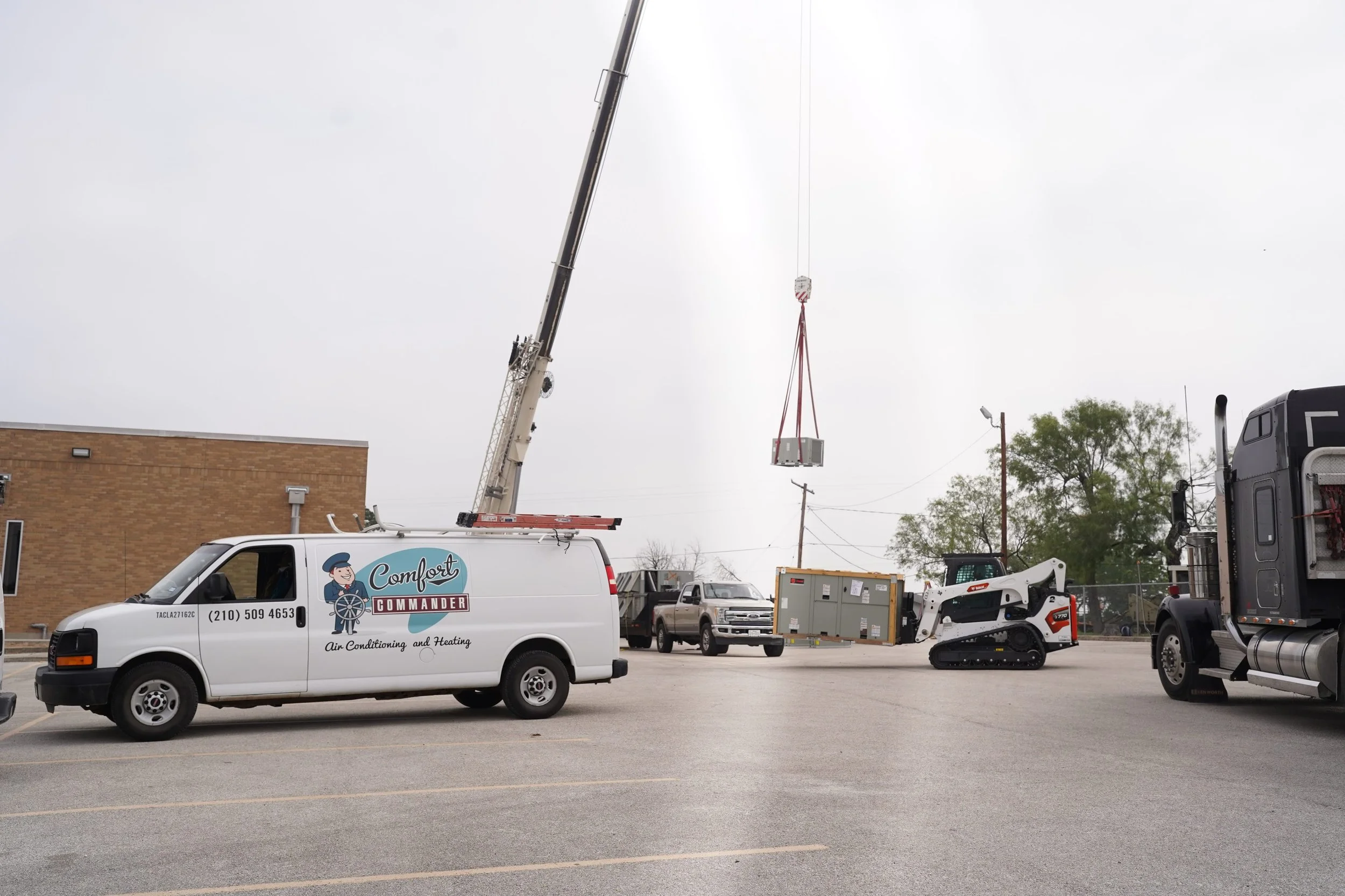 Construction or maintenance equipment in a parking lot, including a crane lifting a container, a compact loader, and utility trucks, with a building and trees in the background.