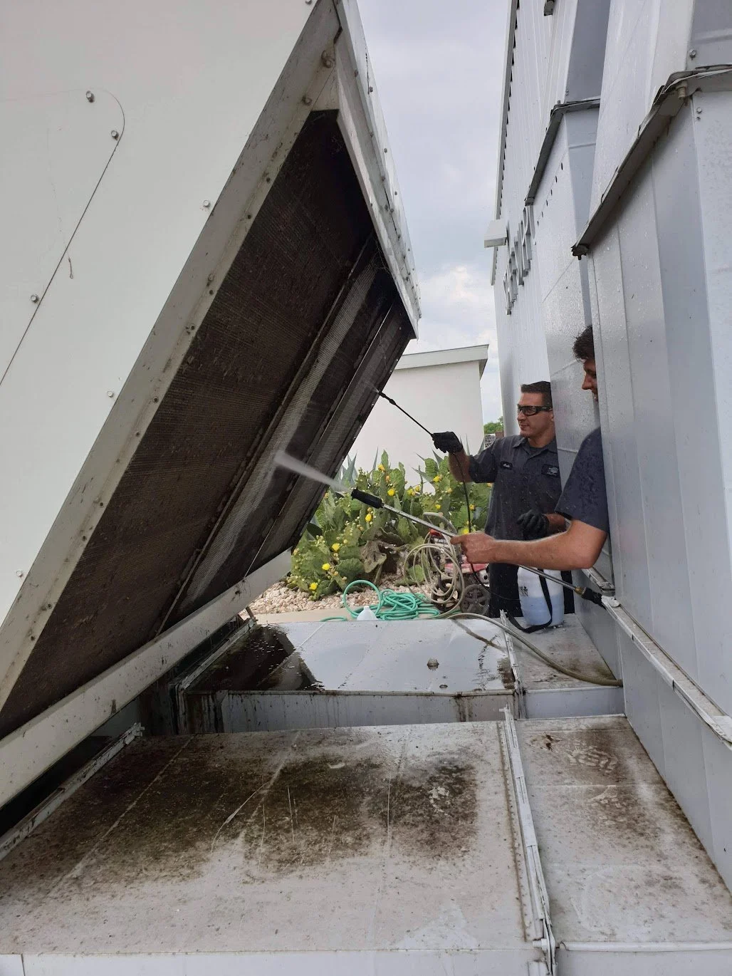 Two men cleaning the exterior of an HVAC unit with pressure washers on a cloudy day.