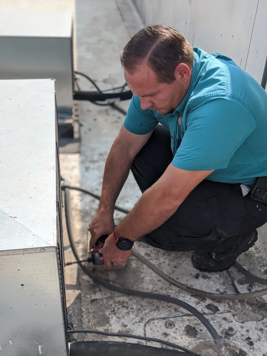 Technician working on an HVAC unit, kneeling and using a power tool on the equipment, with cables and a concrete floor visible.