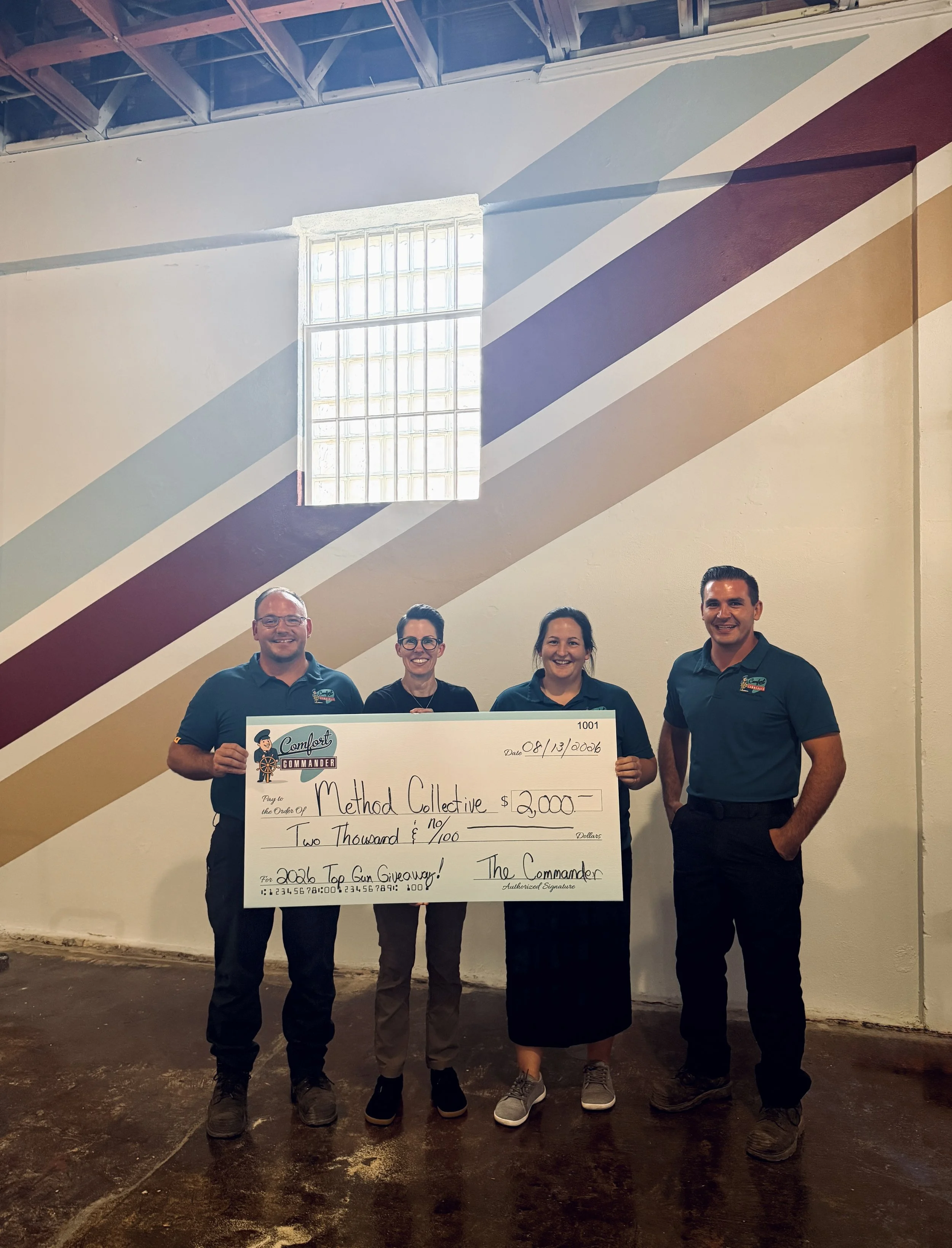 Group of five people standing indoors, holding a large check for $2,000 made out to Method Collective, dated August 13, 2006, in front of a colorful mural wall and a large window, with smiles on their faces.