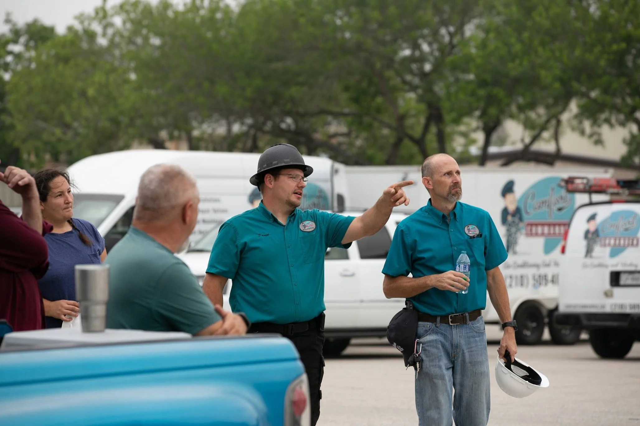 A group of people, including two men in blue shirts and one wearing a helmet, are standing outside near a van with a logo, engaged in a discussion. One of the men in a blue shirt is pointing as he speaks.