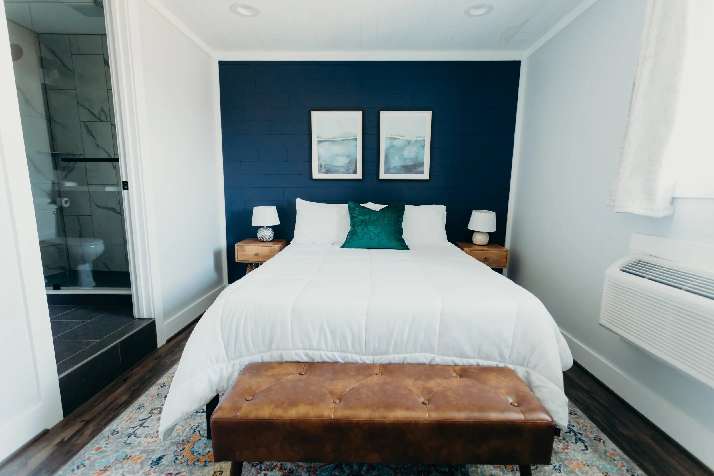Modern boutique hotel room interior with a king bed, crisp white linens, dark blue accent wall, and a wooden bench.