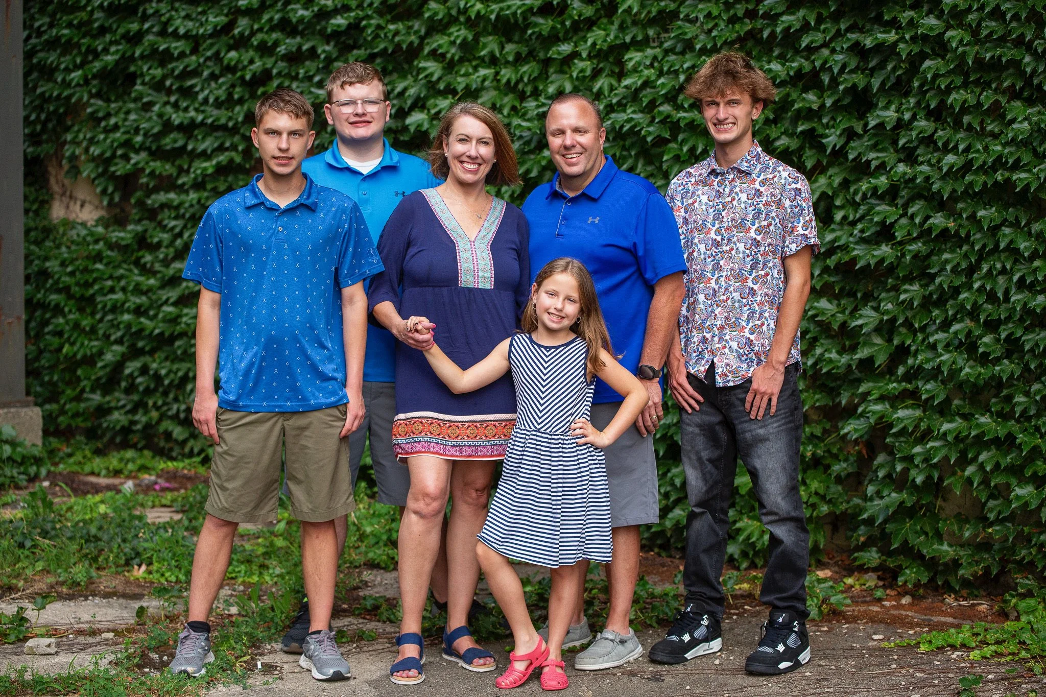 A family of stands outdoors in front of a green leafy wall, smiling at the camera. The group includes three boys,  a woman, and a young girl. The boy on the left wears a blue patterned shirt and khaki shorts, the middle boy wears glasses and a blue polo, and the boy on the right wears a short-sleeved shirt with a floral pattern. The woman in the center has short brown hair and wears a navy blue dress with colorful embroidery. The man next to her wears a blue polo shirt, and the young girl in front wears a blue and white striped dress. They appear happy and close, holding hands and standing closely together.