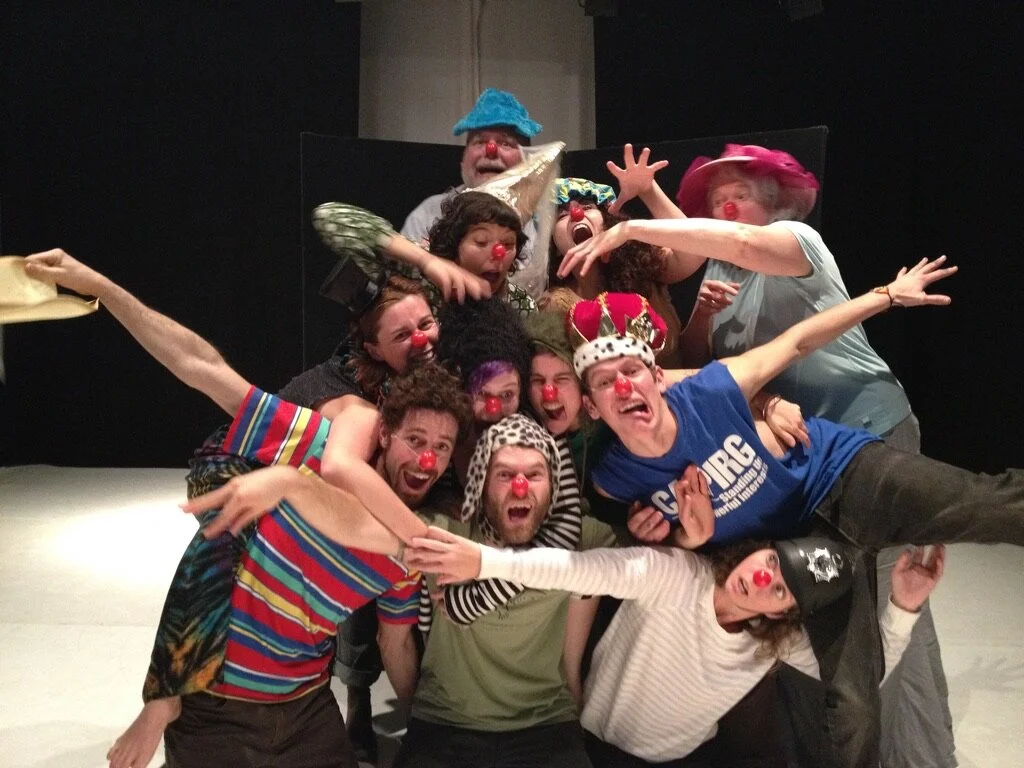 Eleven clowns dogpile in a somewhat triangular formation with many hands outstretched for balance. Facial expressions range from "I'm gunna get you," to "Help I'm falling" to "I'm so happy I could scream" to "Wait, how did I get here?"