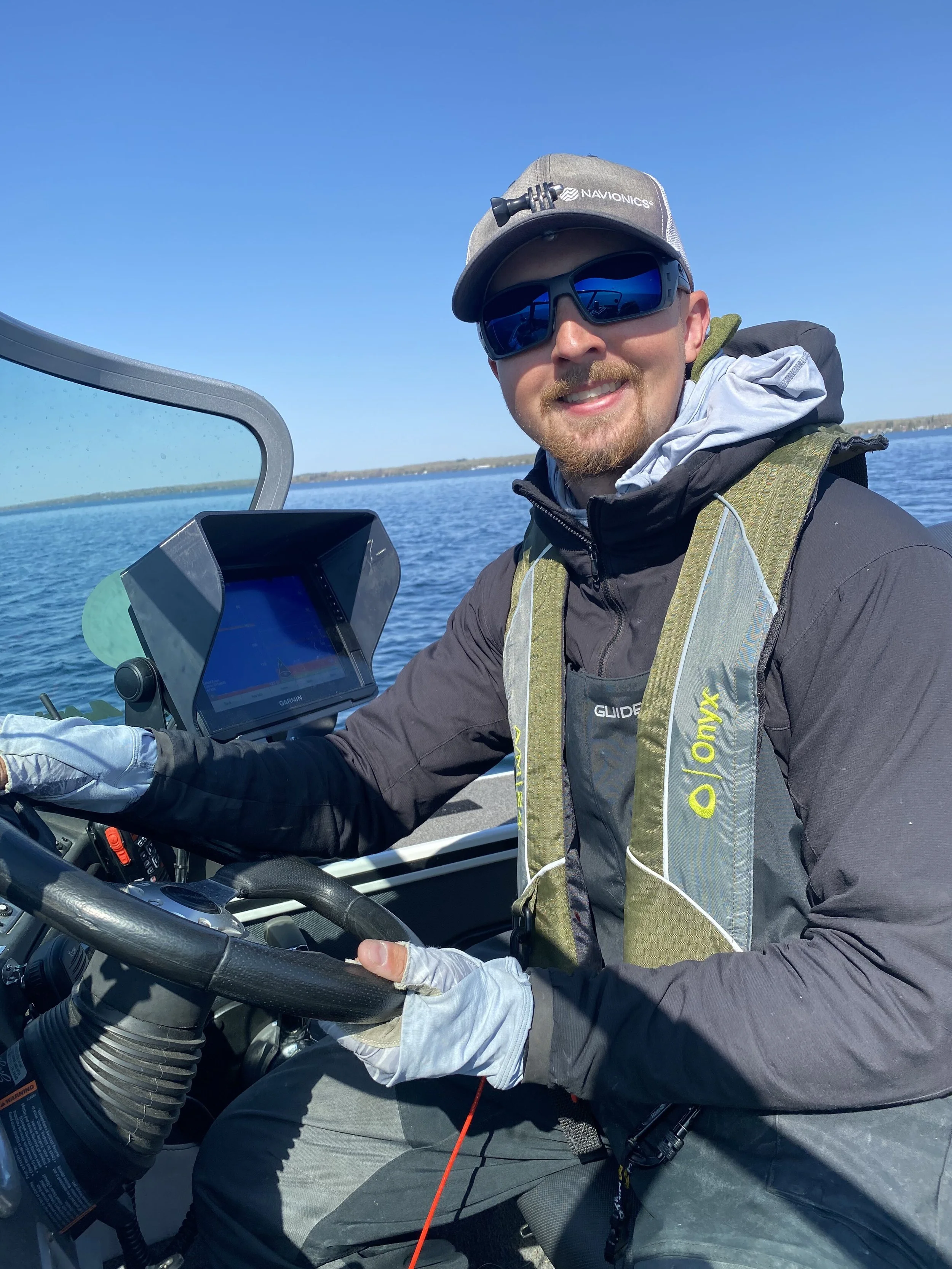 Man in sunglasses smiling while steering a boat on a lake on a sunny day.