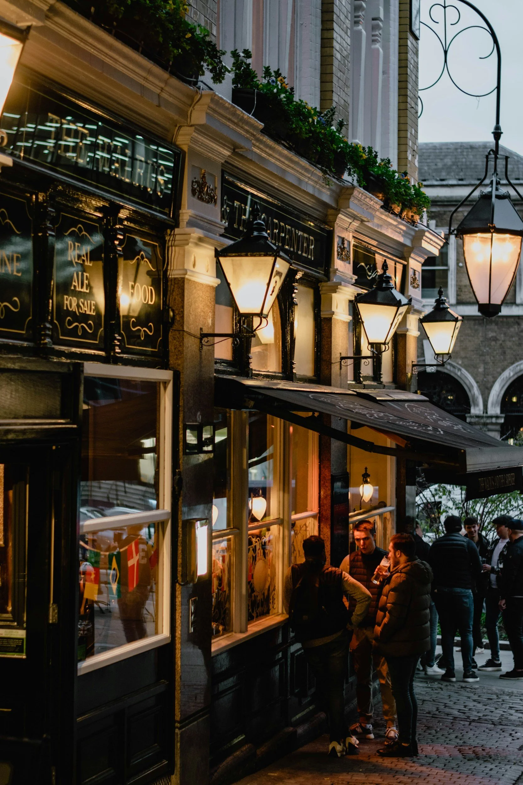 People waiting outside a restaurant or bar in the evening, illuminated by vintage-style lanterns, with a storefront displaying signs for food and beer.
