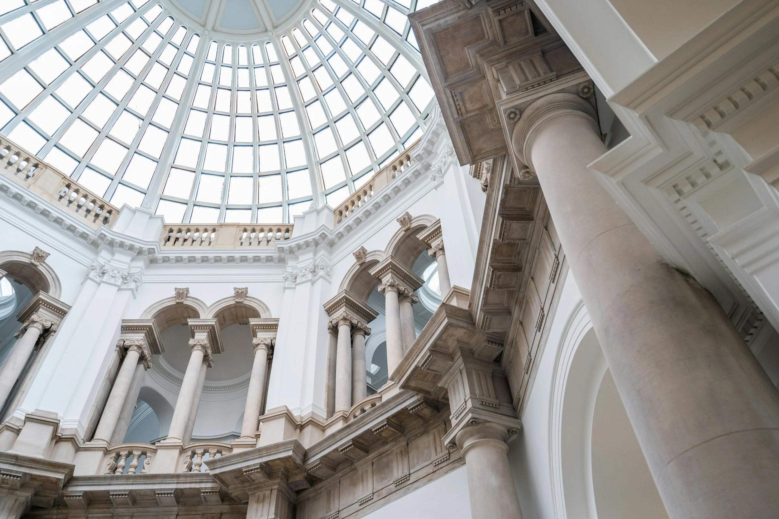 Interior view of a grand building with a domed glass ceiling, white ornate columns, arches, and marble details.