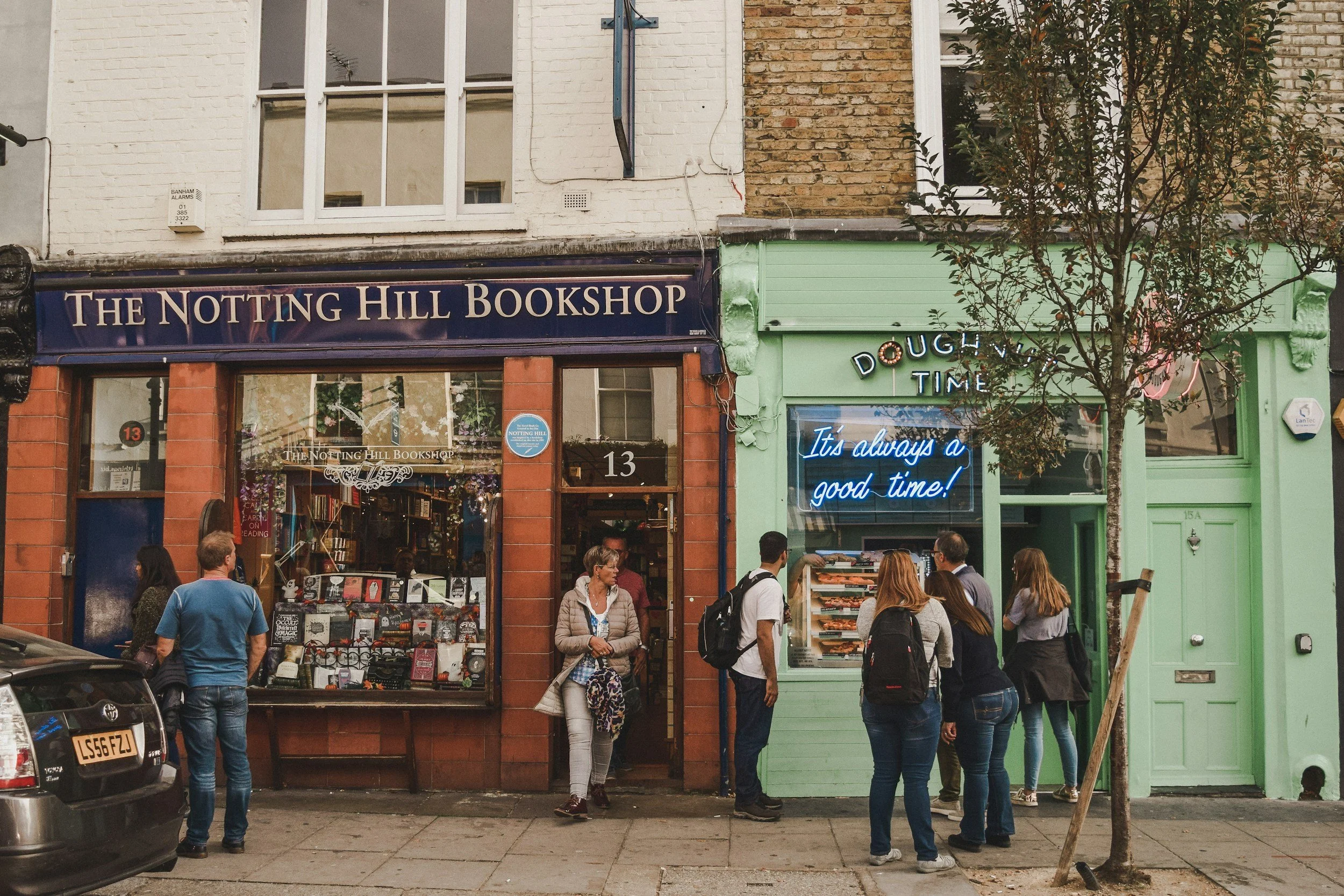 People gathered outside a bookstore named 'The Notting Hill Bookshop' on a busy street with a storefront window displaying books and a neon sign that reads 'It's always a good time!' next to a green storefront, with a small tree in front.