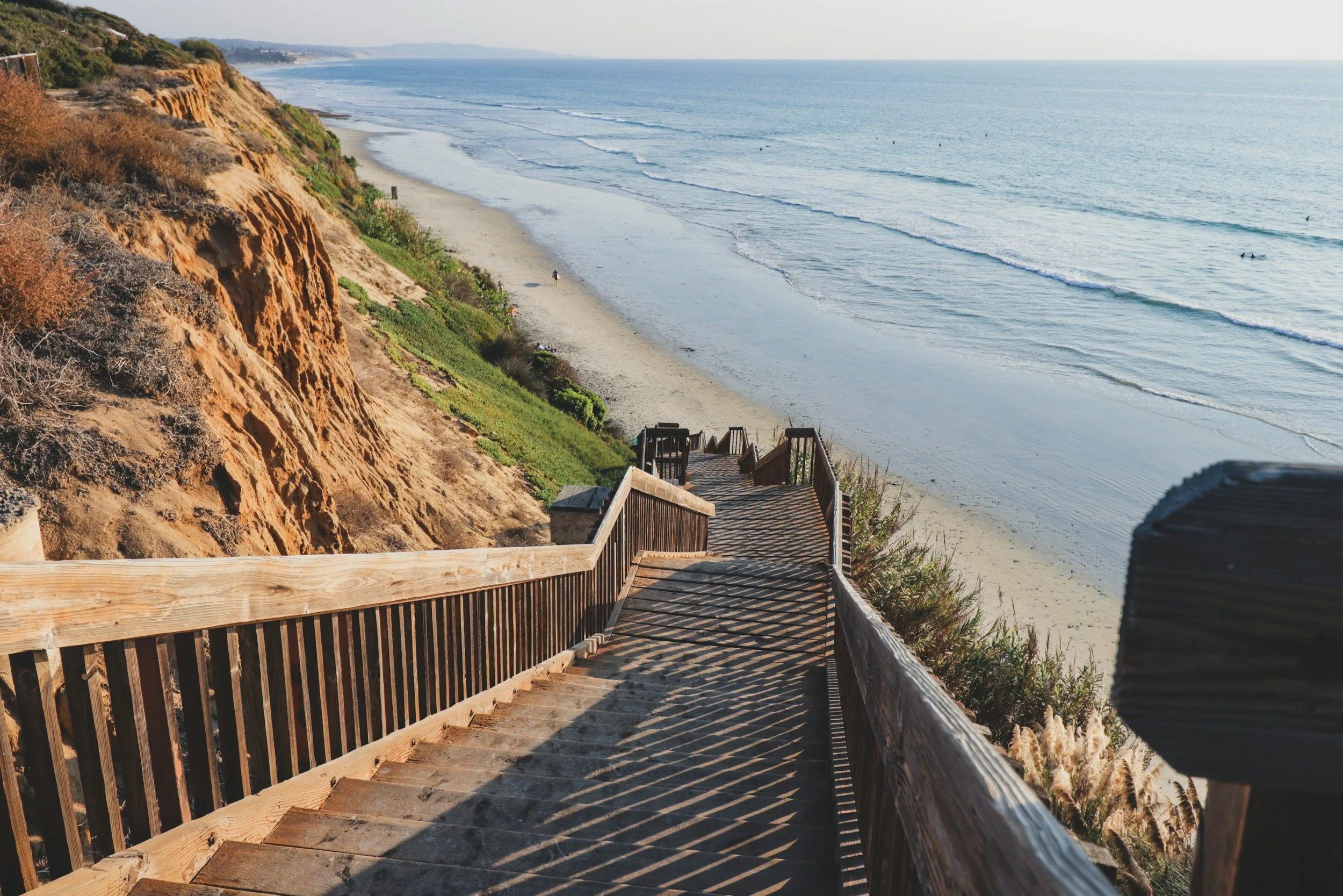 Wooden staircase leading down to a sandy beach with cliffs on the left and ocean waves on the right. Few people walking on the beach, with the ocean and distant land in the background.