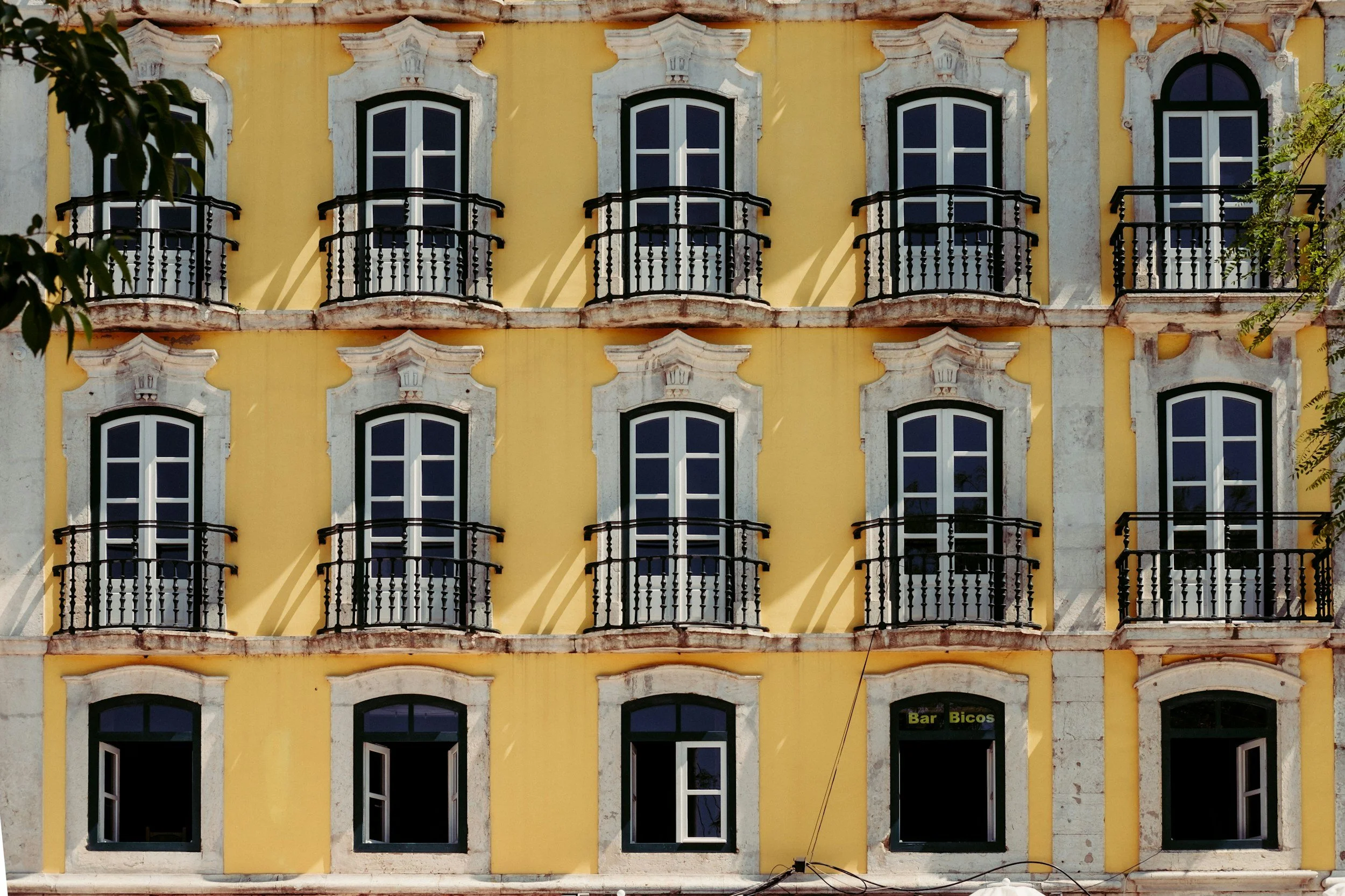 Yellow building with multiple balconies and white-framed windows, some open, and a sign at the bottom reading 'Bar Bicos'.
