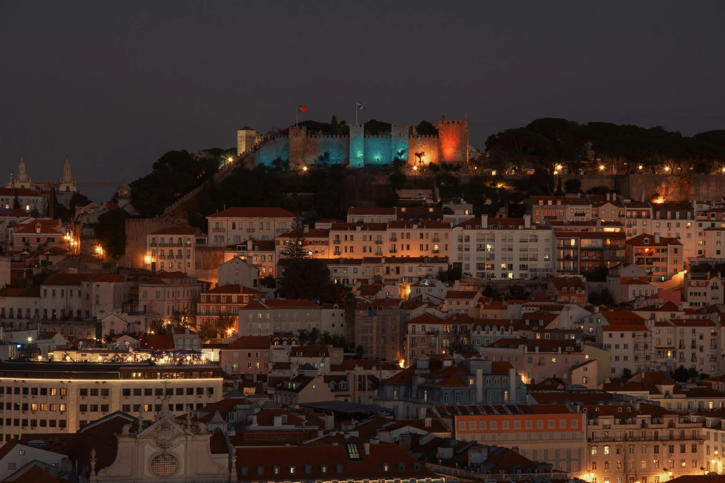 Night view of a hillside castle in Lisbon, Portugal, illuminated with red, blue, and green lights, overlooking a cityscape with residential buildings and some church steeples in the distance.