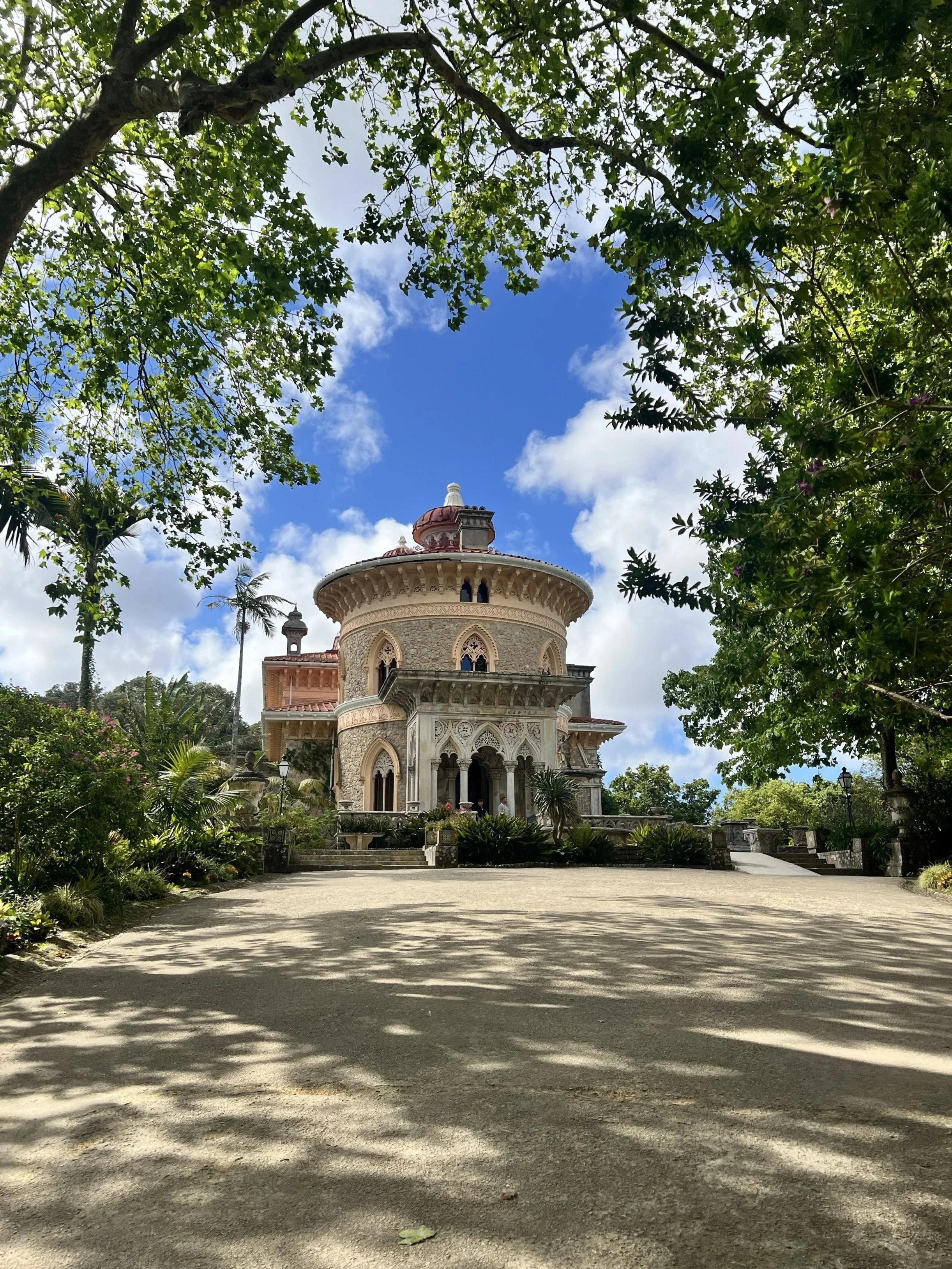 Monserrate Palace in Sintra, Portugal featuring its architectural details, surrounded by lush greenery and trees, under a partly cloudy blue sky.