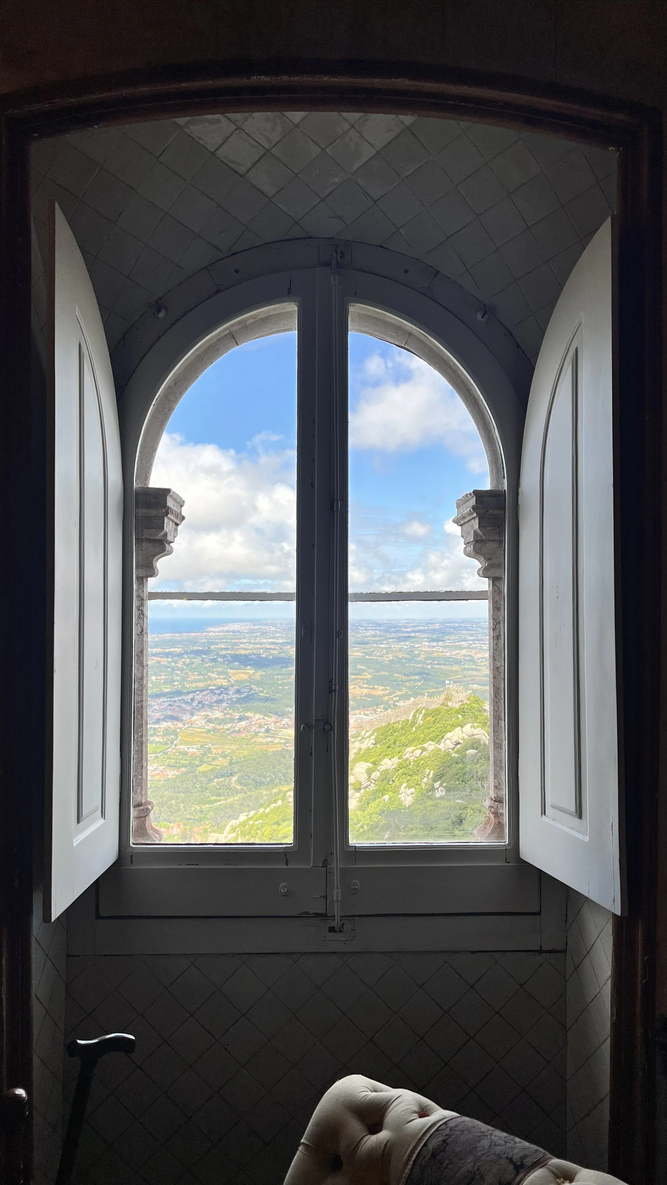A view through a large arched window in Pena Palace in Sintra, Portgual, with open white shutters, revealing a scenic landscape with hills, fields, and a partly cloudy sky.
