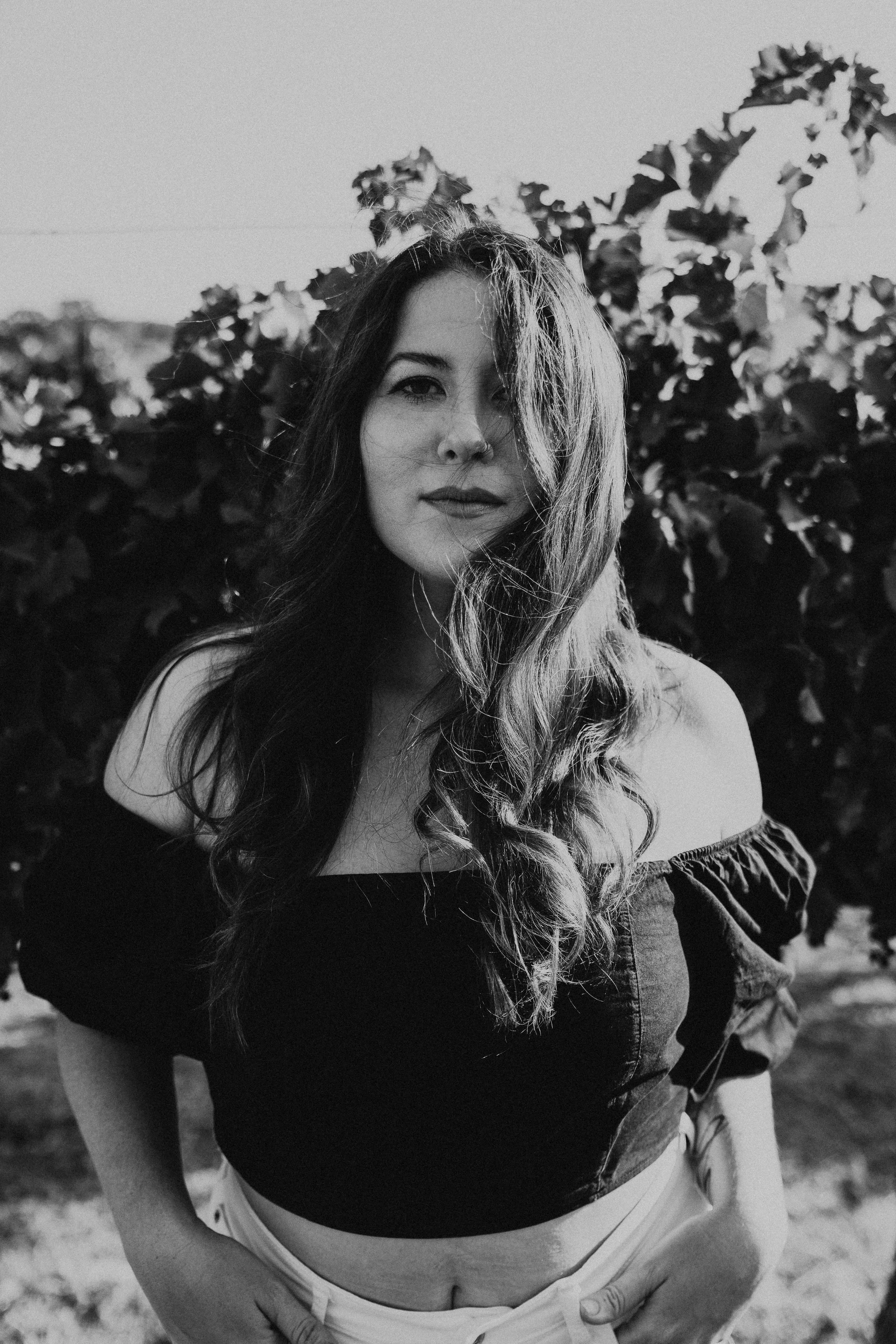 A black-and-white photo of a woman with long hair, wearing a short-sleeved top with ruffled sleeves and white pants, standing outdoors with foliage in the background.