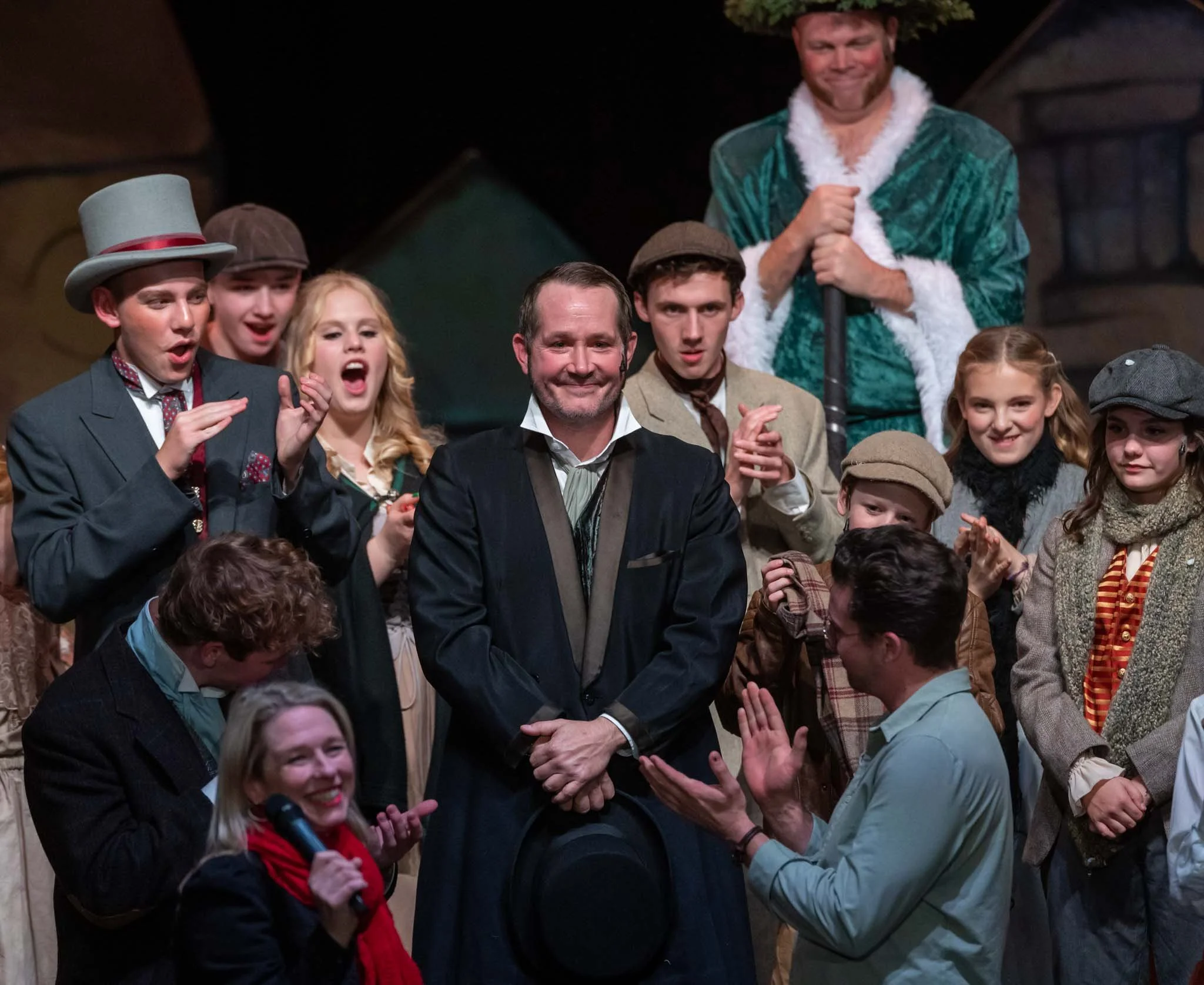 Group of people in theatrical costumes on stage, some clapping and smiling, with an adult man in the center wearing a suit. A person dressed as Santa Claus is in the background.