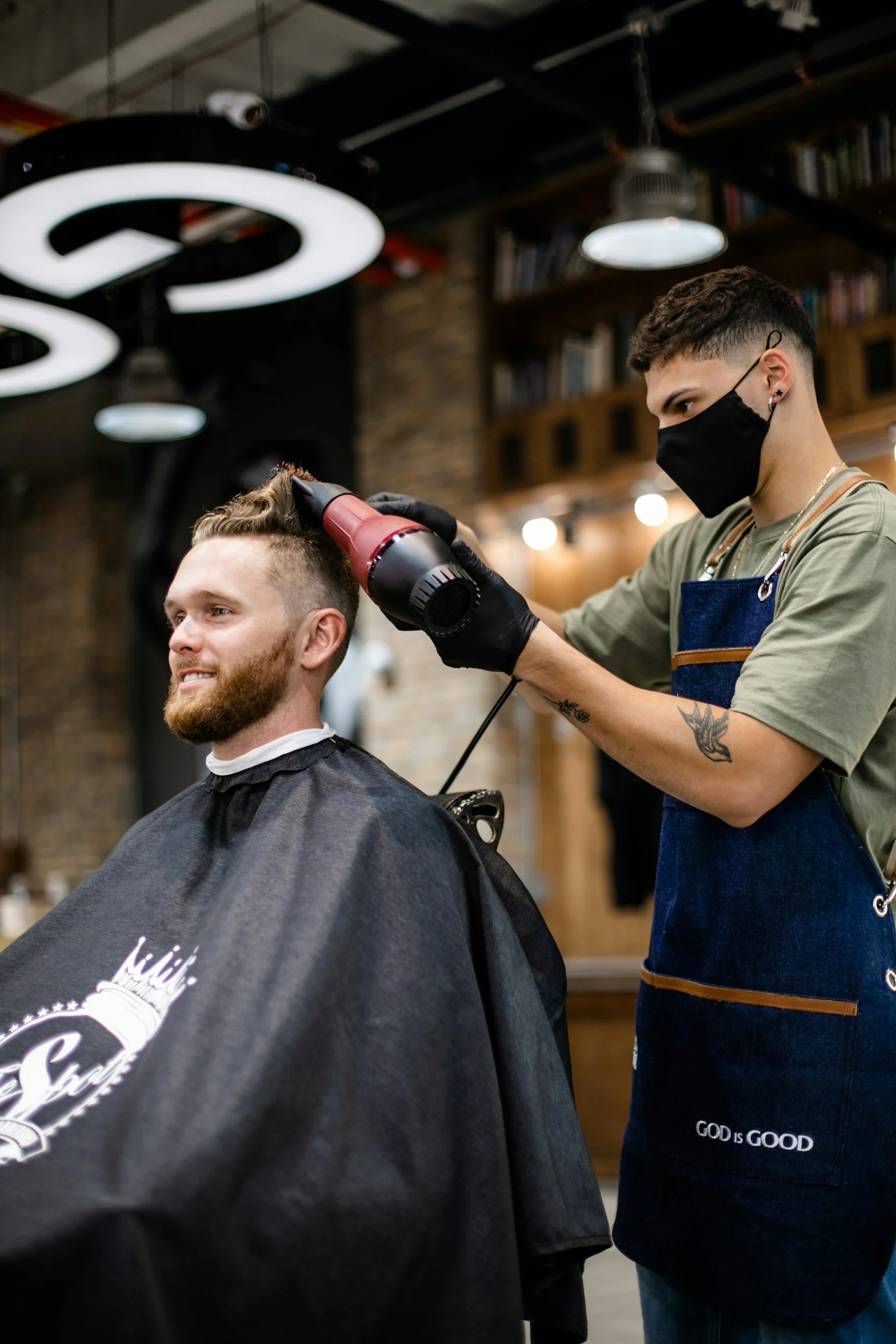 Barber giving a haircut to a male customer in a modern barber shop, with a bookshelf on the wall and hanging lights overhead.
