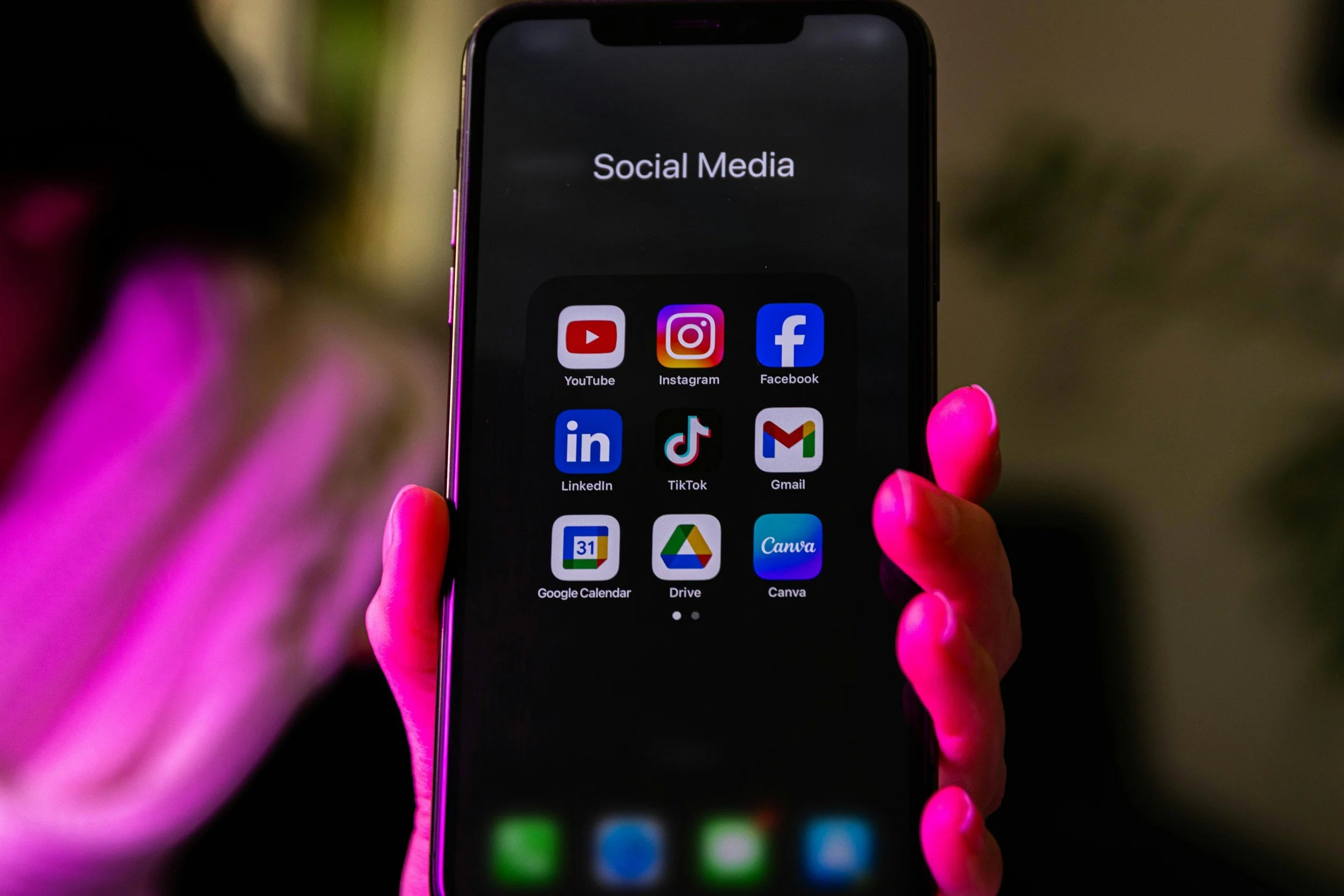 Person with pink nails holding a smartphone displaying social media app icons, with a dark background.