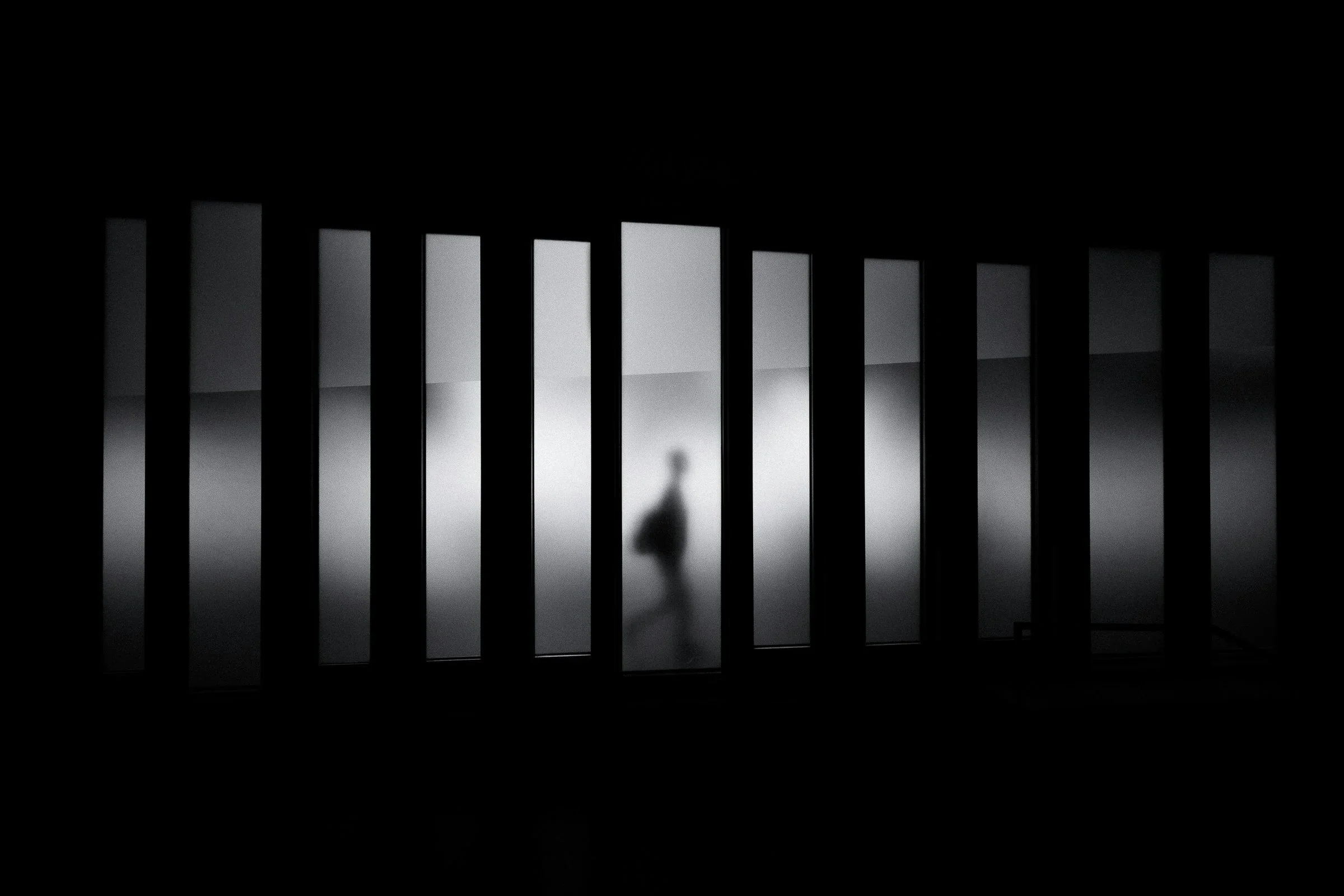A blurred silhouette of a person walking past vertical black bars with a gray background.