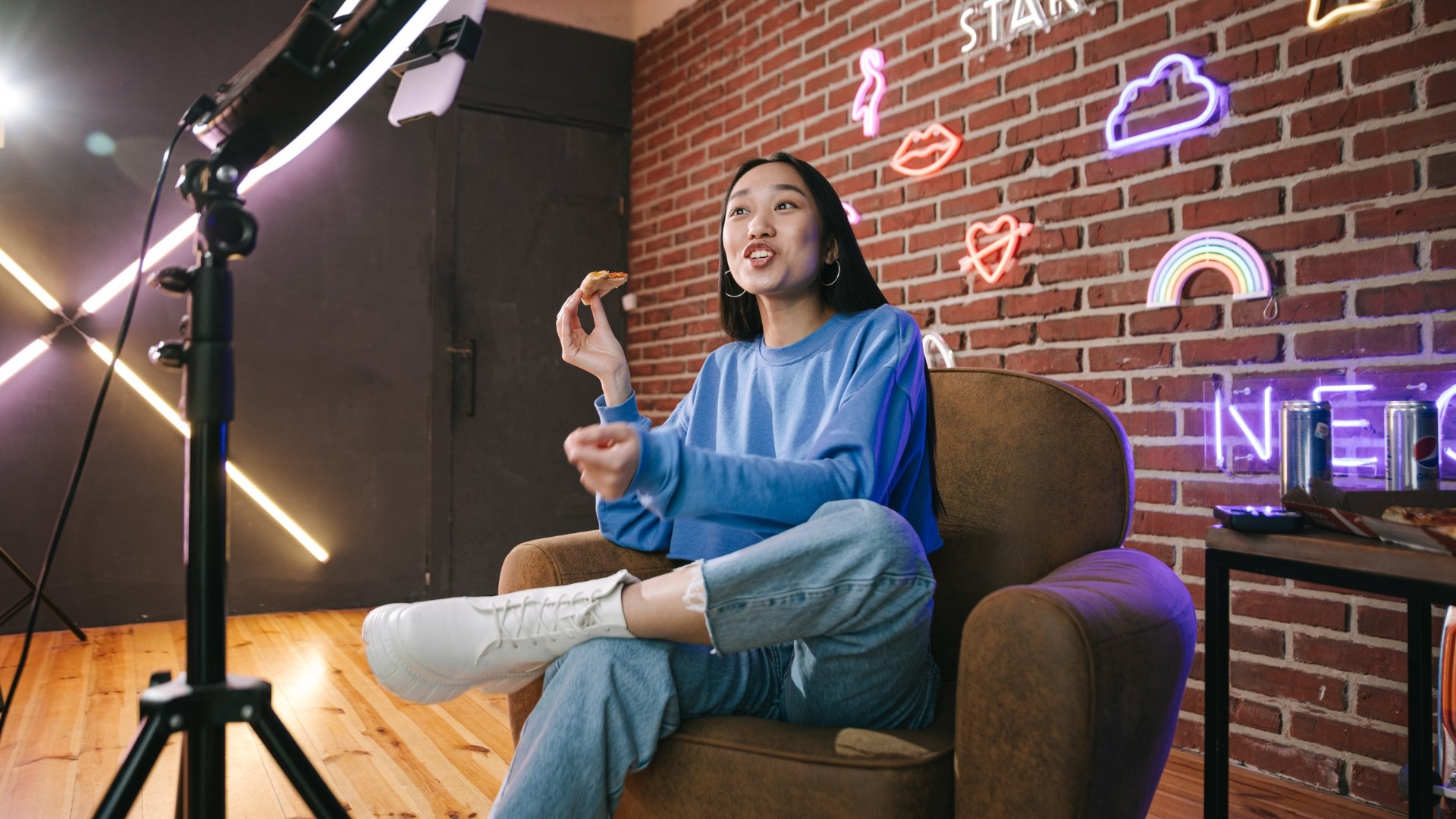 A young woman in a blue sweatshirt and ripped jeans sitting on a brown couch, holding a slice of pizza, in a room with exposed brick wall decorated with neon signs including rainbows, hearts, lips, and clouds, with a camera on a tripod recording her.