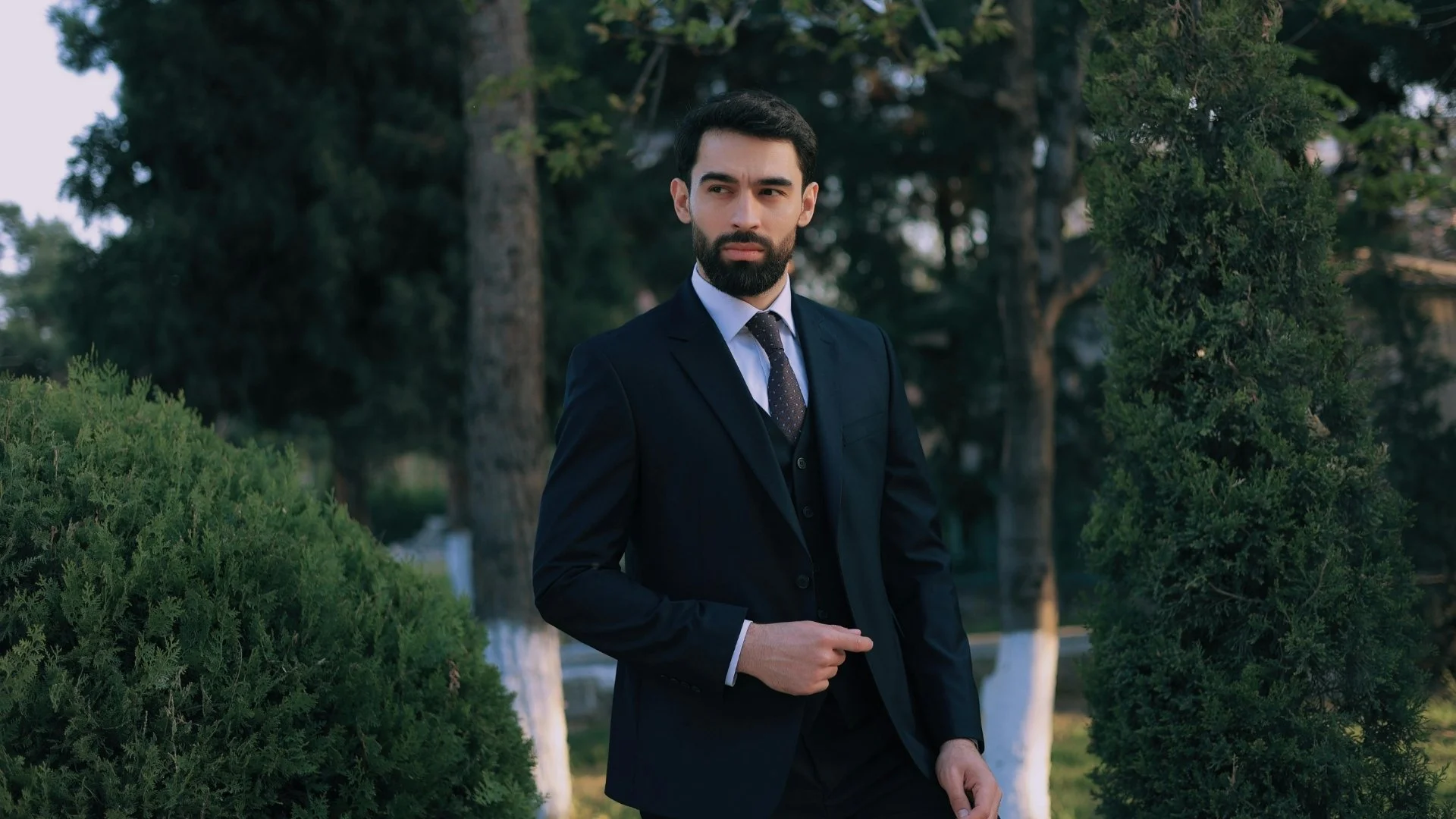 A man in a dark suit and tie standing outdoors amidst greenery, with a serious expression, looking to his left.
