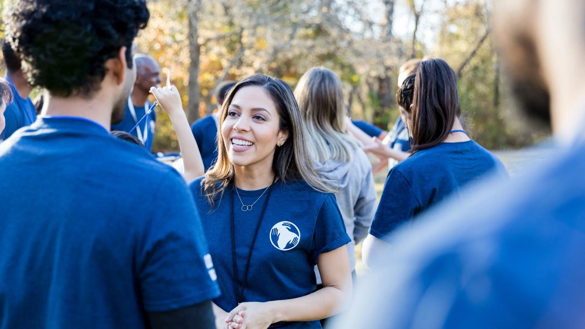 Group of people outdoors in a park, engaging in a discussion or event, with a woman in a blue t-shirt smiling and talking, surrounded by others in casual clothing.
