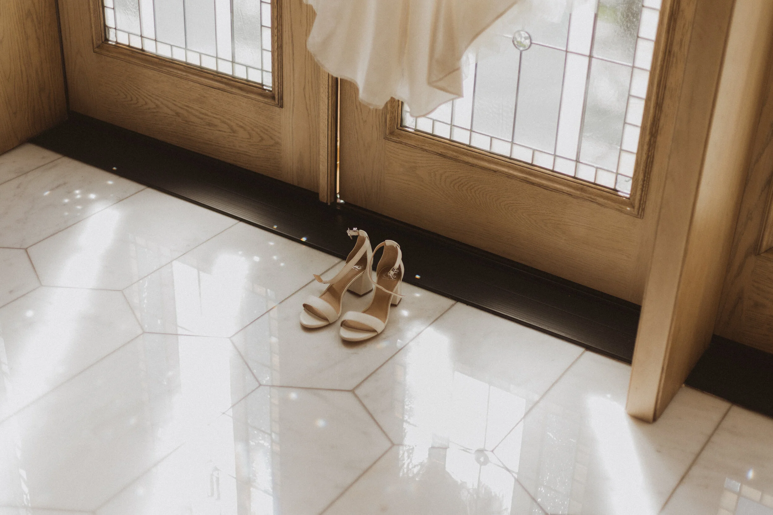 Pair of beige high-heeled sandals placed on a shiny tile floor near a wooden door with glass panels.
