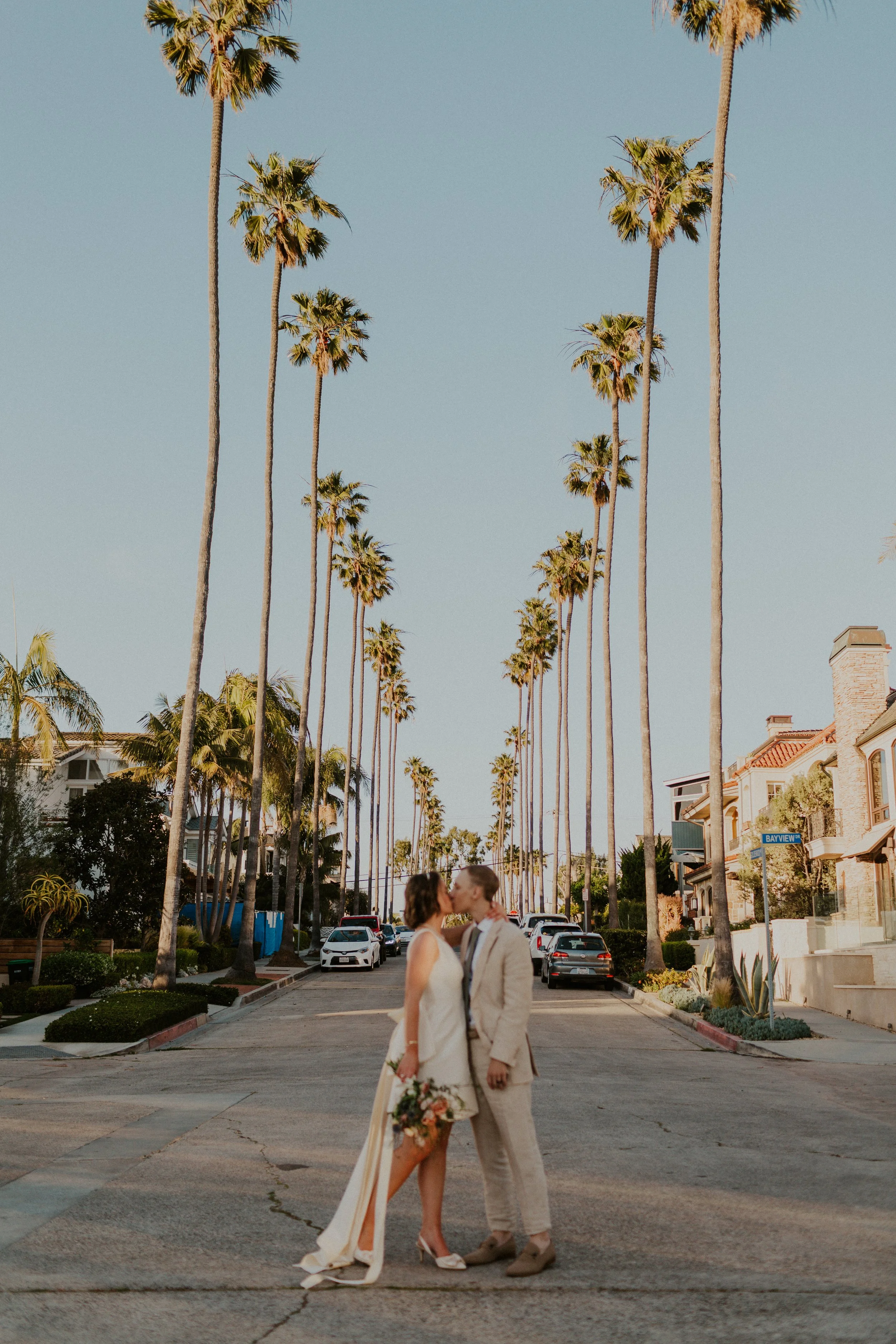 A couple dressed in wedding attire sharing a kiss on a street lined with tall palm trees, with houses and parked cars in the background.