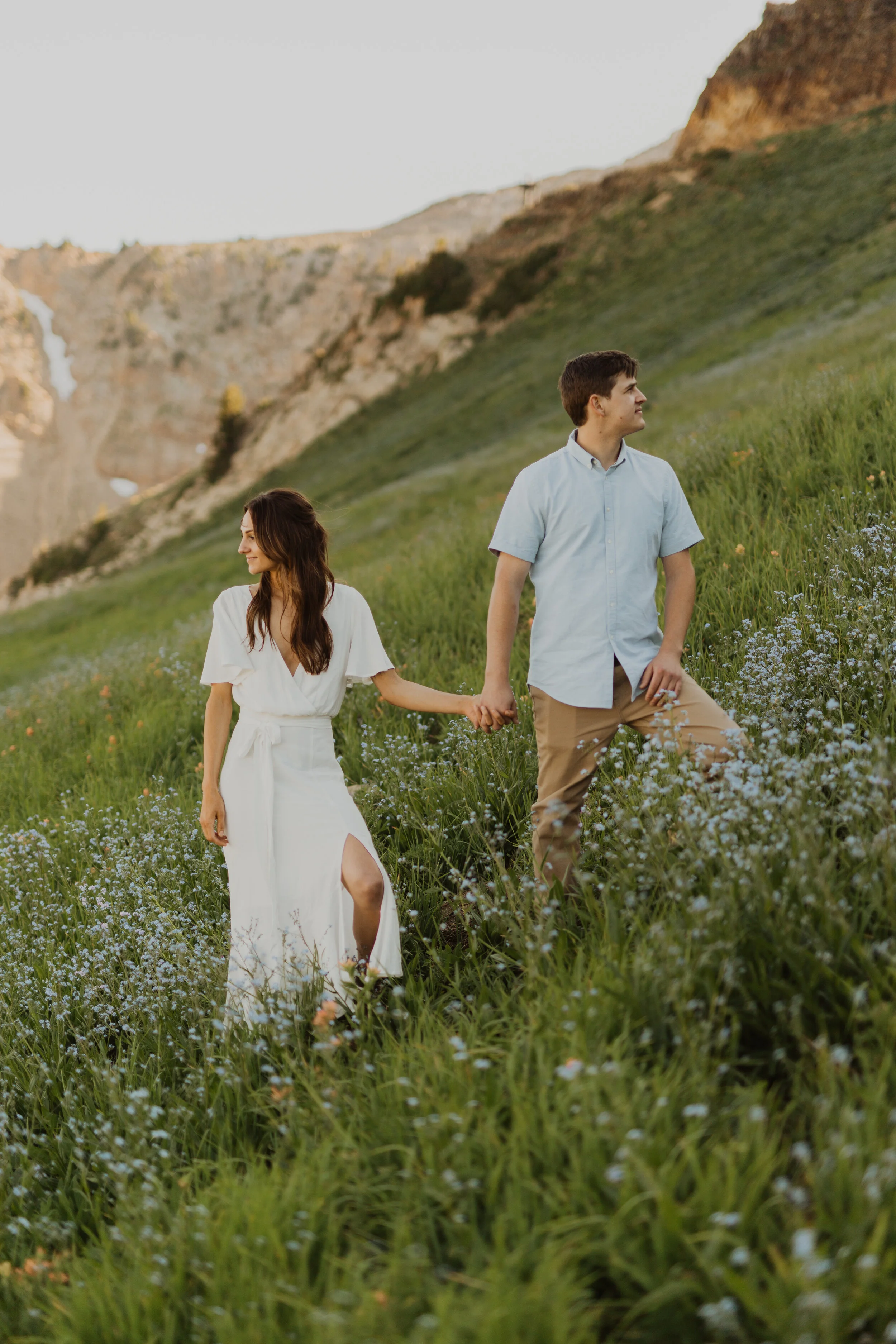 A young man and woman holding hands in a lush green field with wildflowers, surrounded by mountains under a soft sky.