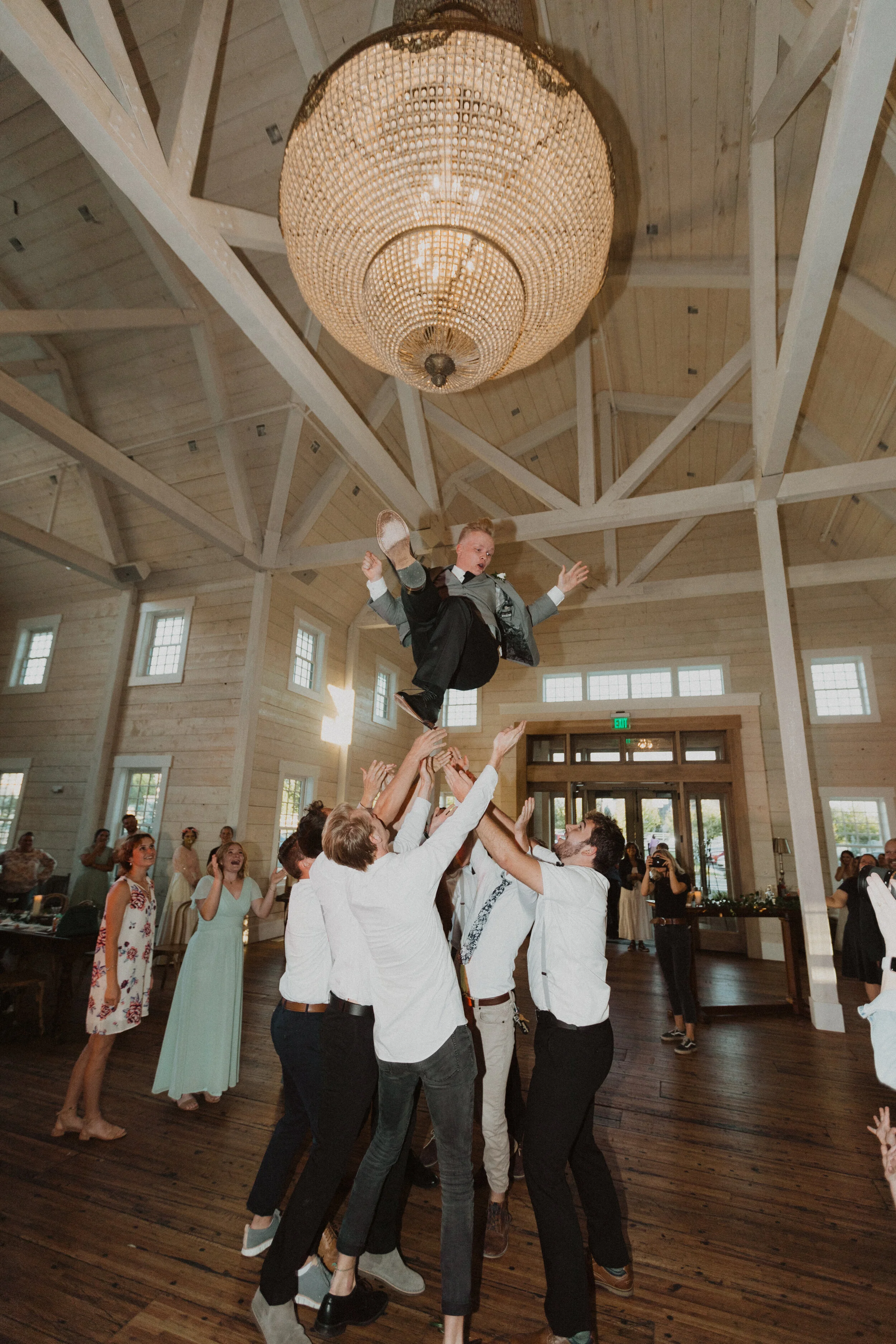 A group of people at a wedding reception lifting a young boy dressed in a suit and tie into the air during a toast or celebration in a rustic wooden hall with high ceilings and a large chandelier.