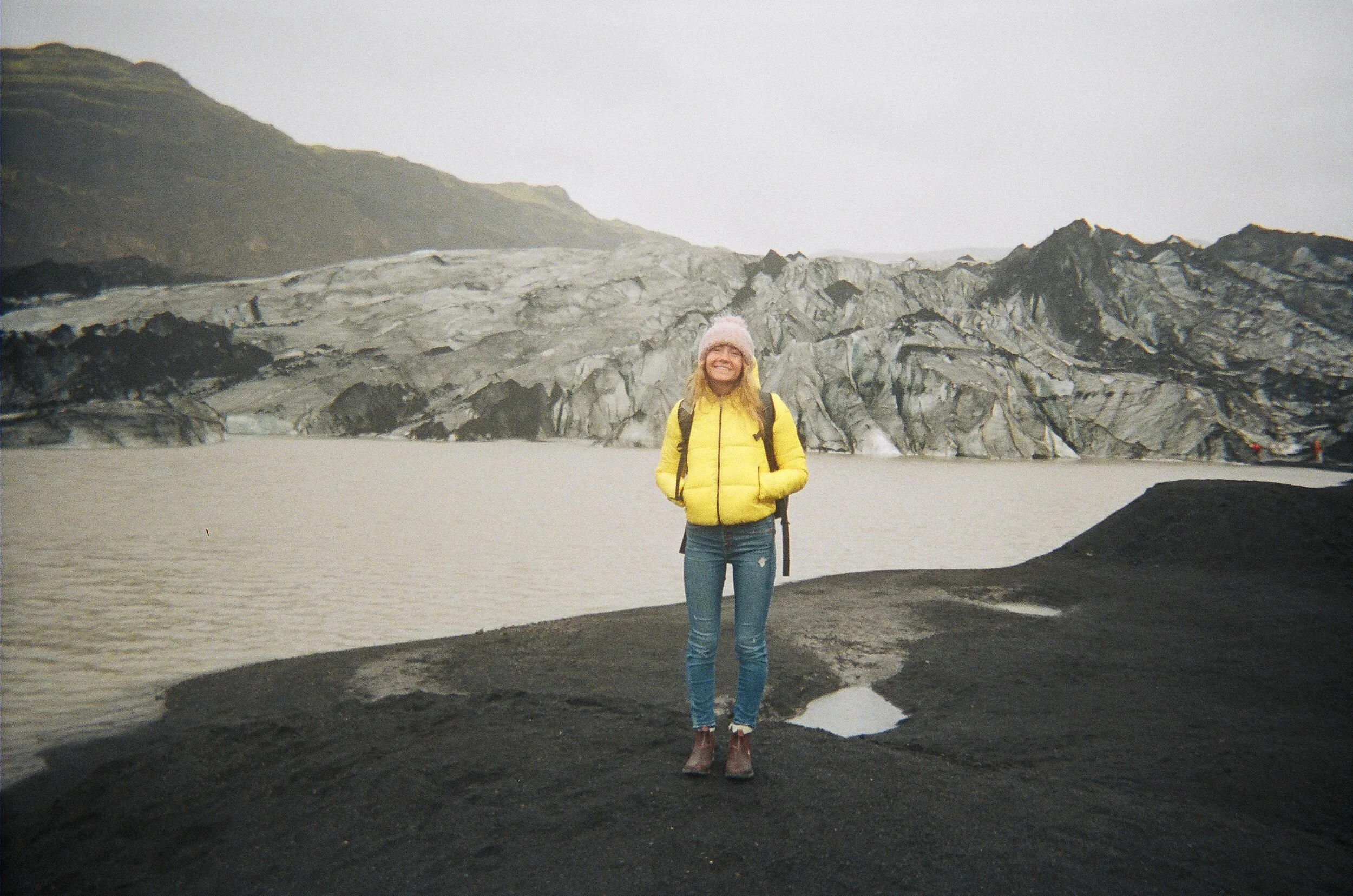 Woman standing on volcanic landscape with glacier and mountains in the background, wearing a yellow jacket, pink hat, and carrying a backpack.