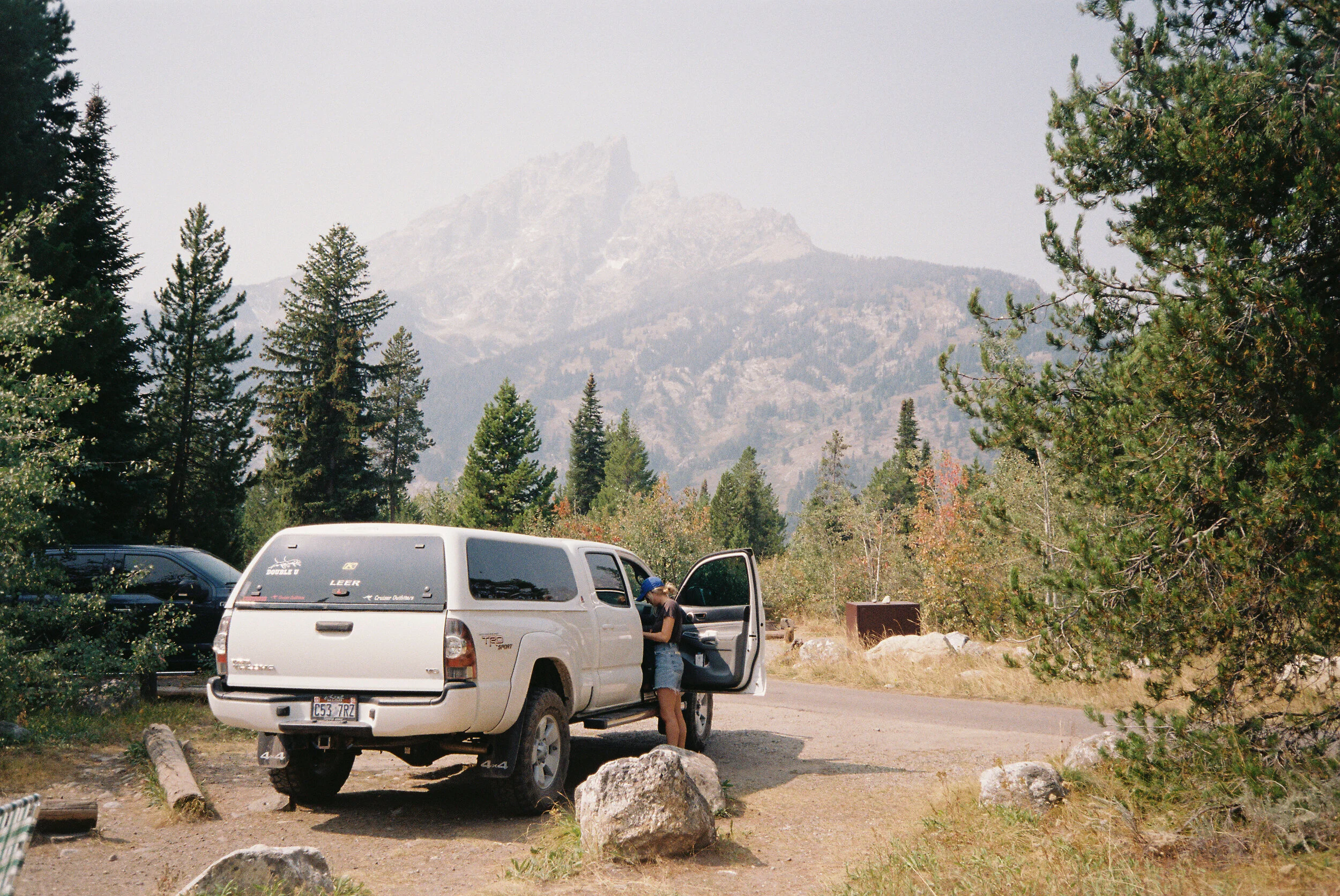 A woman accessing a white SUV parked outdoors with a forested mountain in the background.
