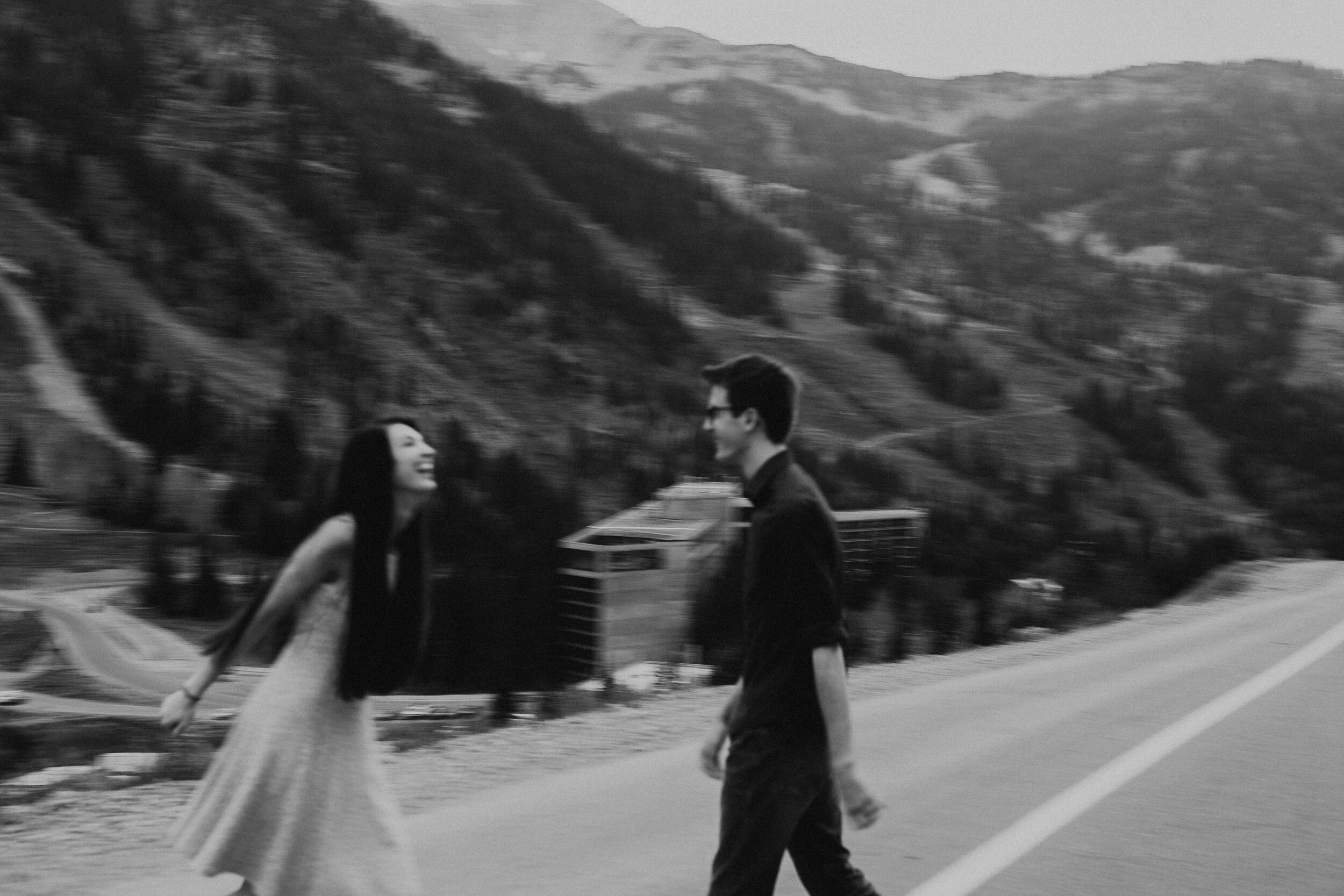A black and white photo of a man and woman holding hands and walking outdoors with mountains in the background.