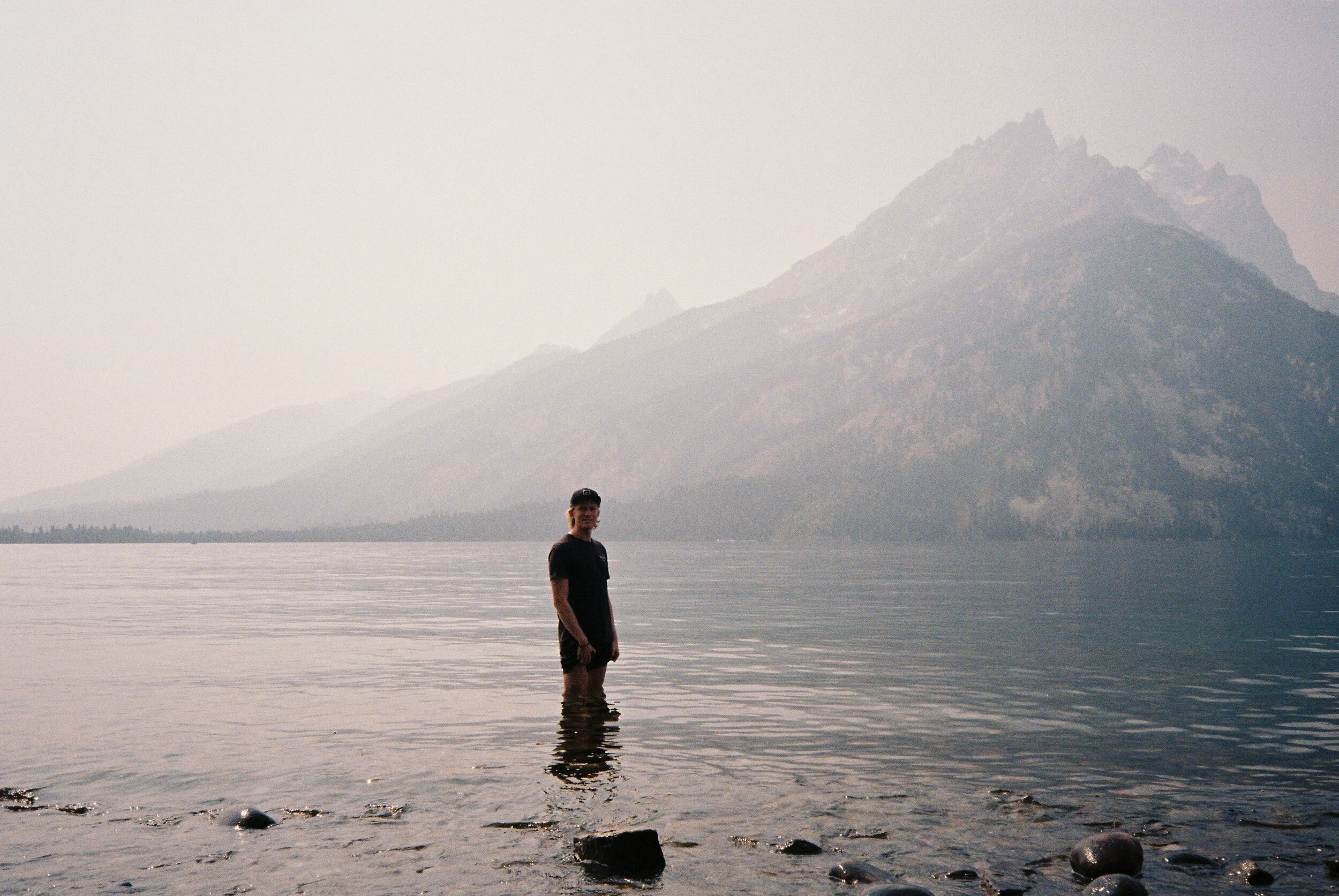 A person standing in a lake with a mountainous landscape in the background, overcast sky.