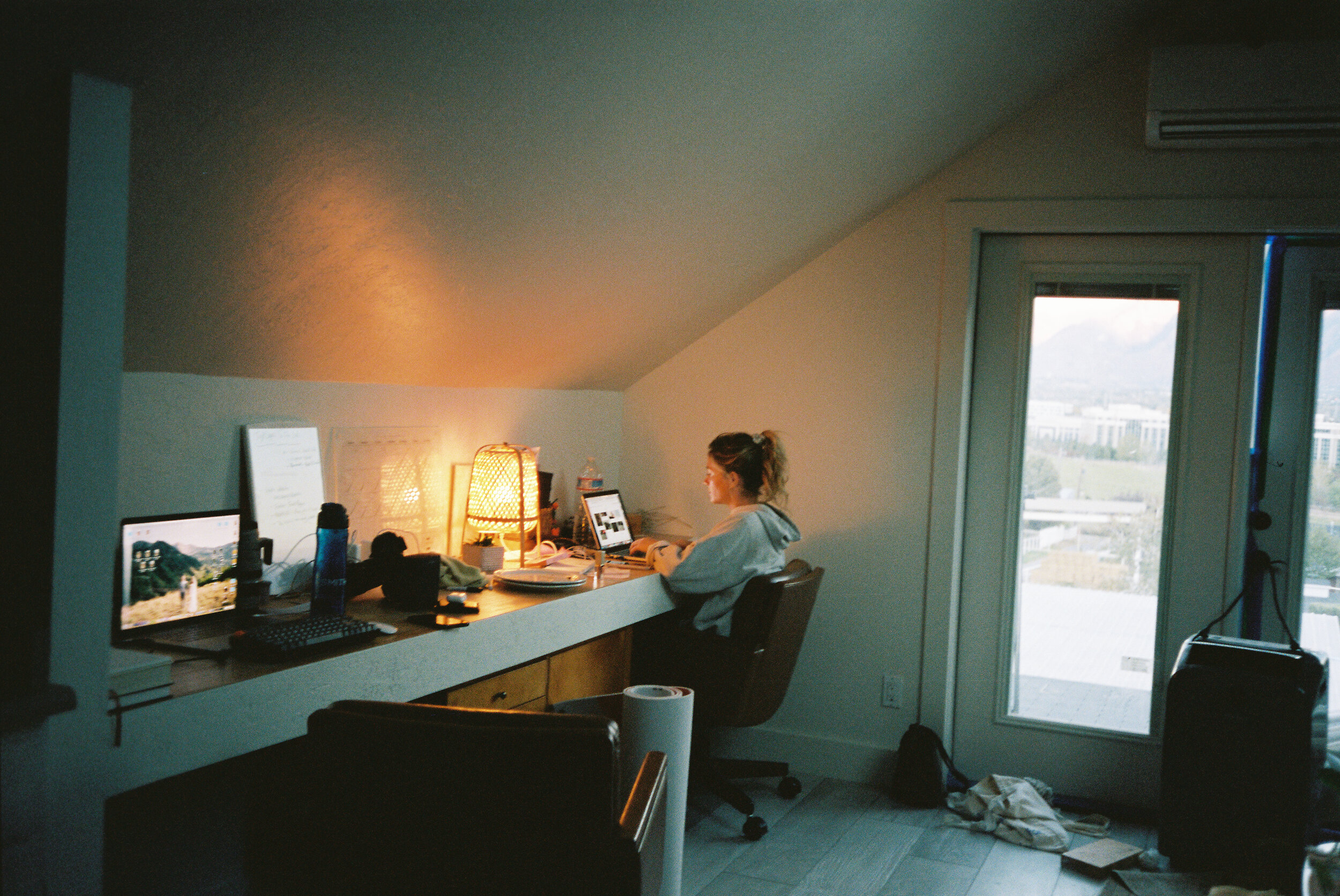 A woman sitting at a desk working on a computer in a room with a sloped ceiling and a large window showing a cityscape.