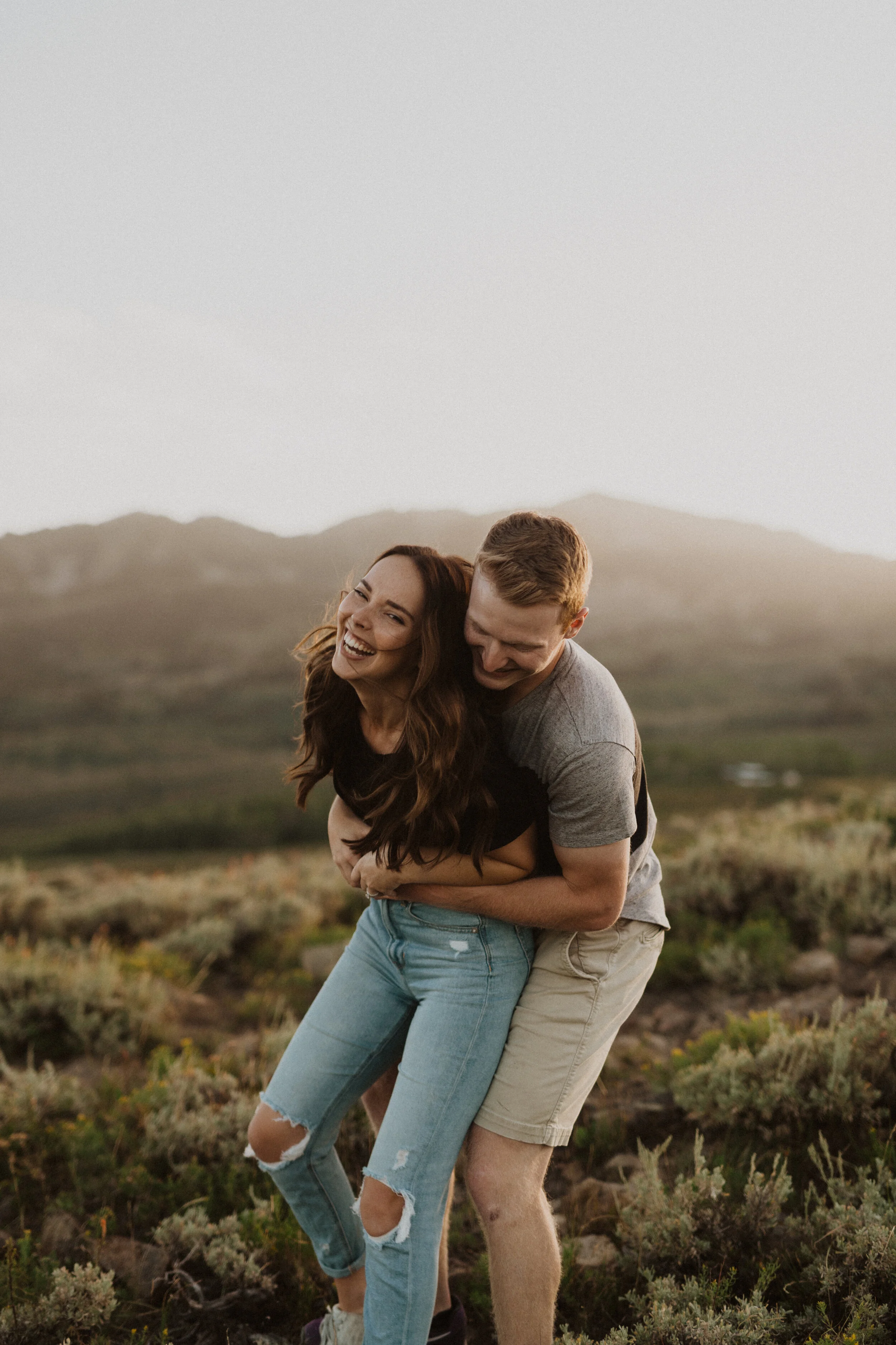 A smiling woman and man are having fun and hugging outdoors in a scenic landscape during sunset, with mountains in the background.