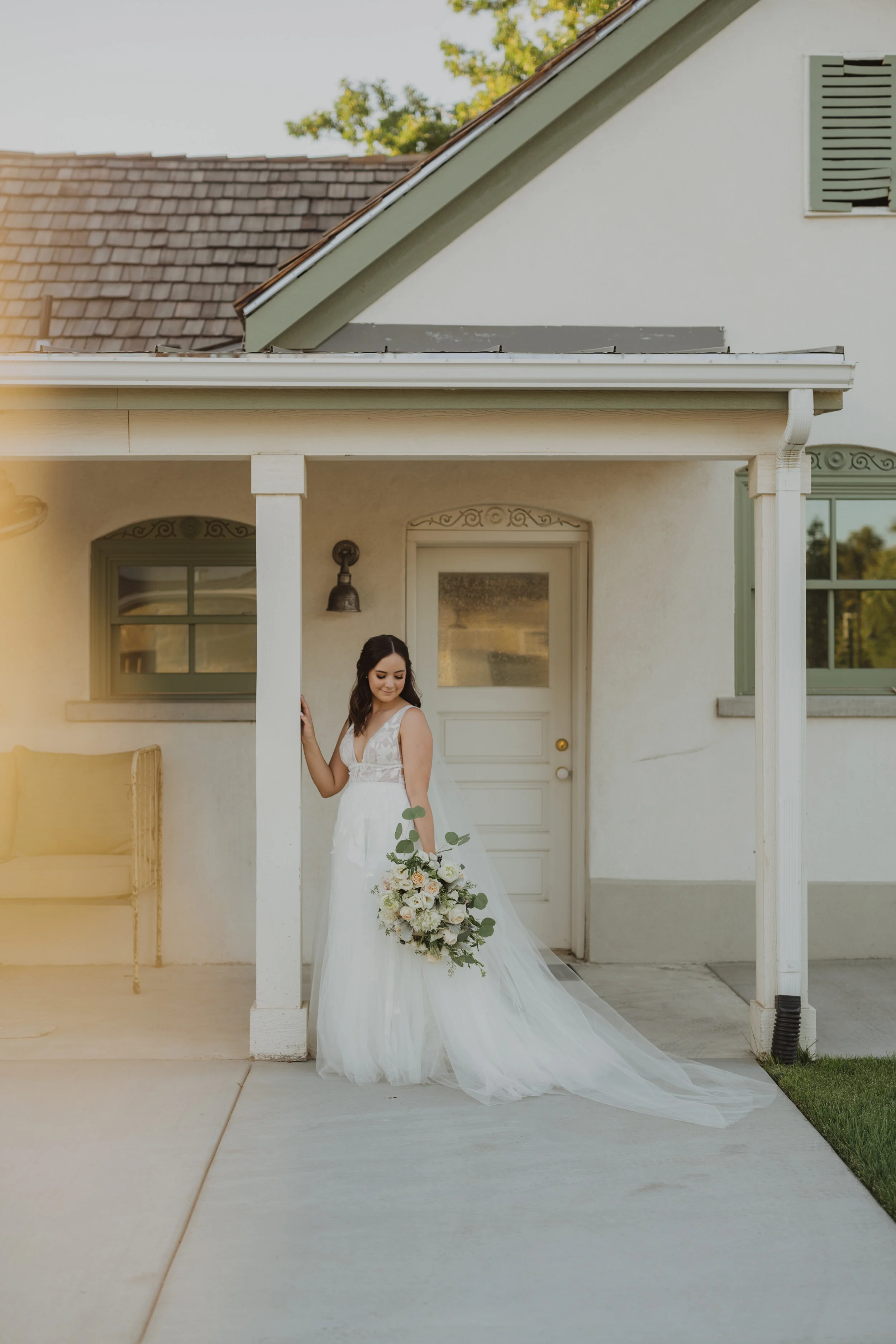 A bride in a white wedding gown holding a large bouquet of white and blush flowers with greenery, standing on a porch of a white house with green window frames, a gray roof, and a small window with shutters. She is smiling and looking down, with her 