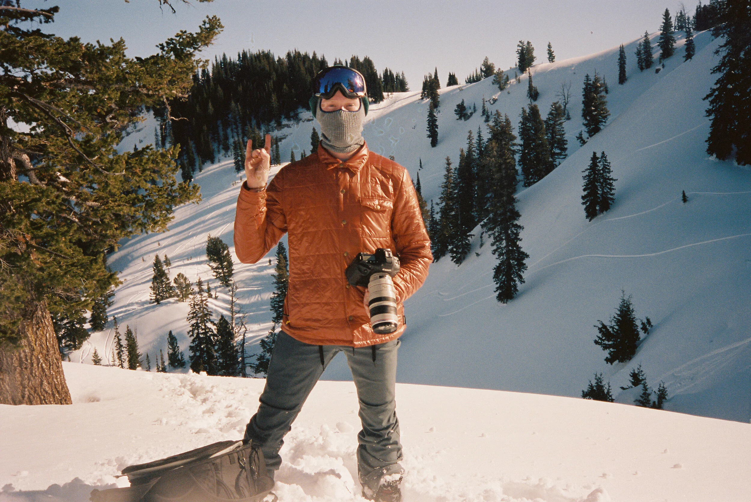 A person wearing a beige face covering, goggles, a brown jacket, and gray pants standing in a snowy mountain landscape, holding a camera, with trees and snow-covered slopes in the background.