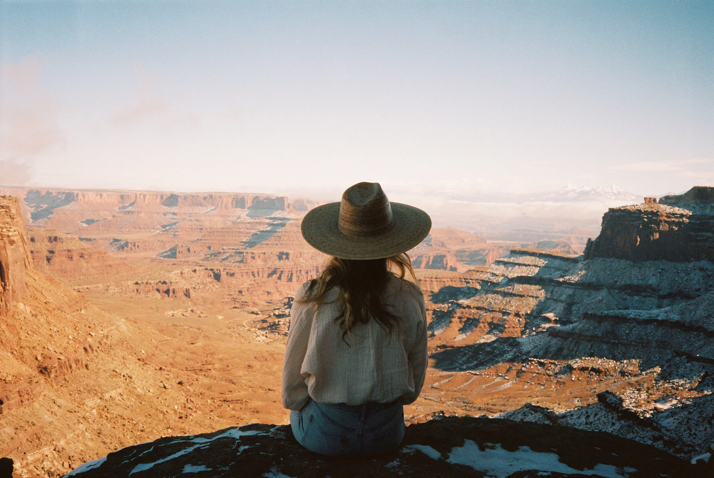 A woman with long hair wearing a wide-brimmed hat and casual clothes, sitting on a rock and overlooking the Grand Canyon.