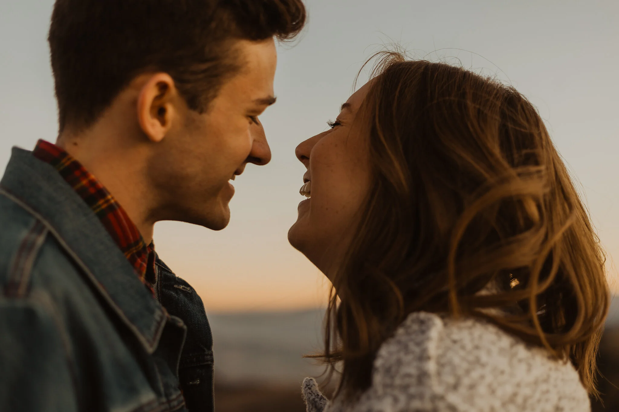 A man and a woman are close to each other, smiling, with their foreheads touching against a background of a sunset or sunrise.