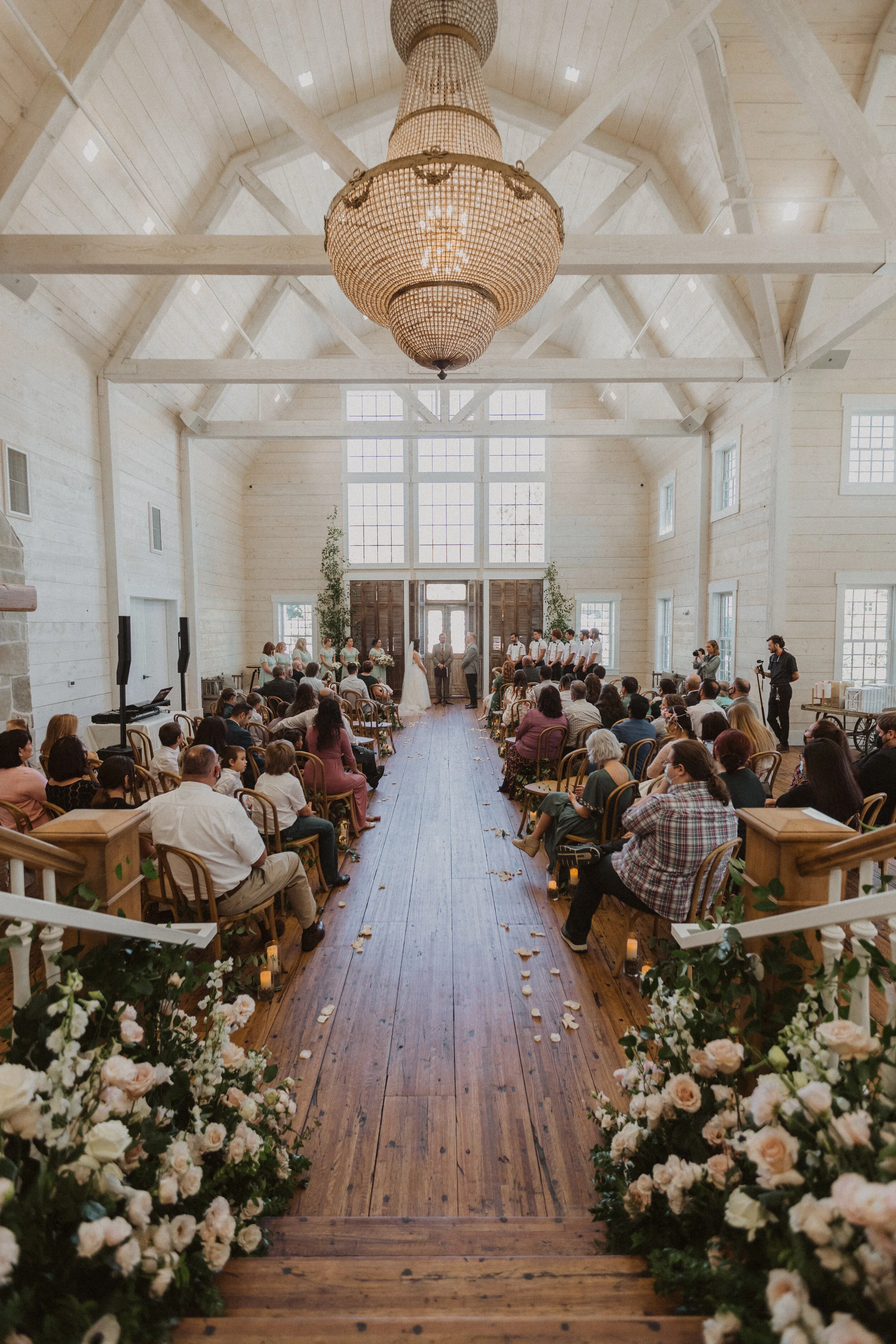 A wedding ceremony inside a large, bright, wooden chapel with high ceilings and a central chandelier. Guests sit on either side of a wooden aisle decorated with flowers and candles, facing a couple at the front with an officiant and bridesmaids and g